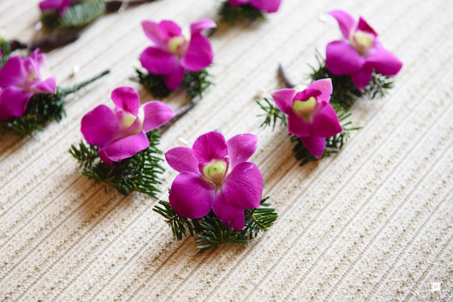 Pink orchid boutonnieres with green fern accents arranged on a woven white surface for a wedding or special event.
