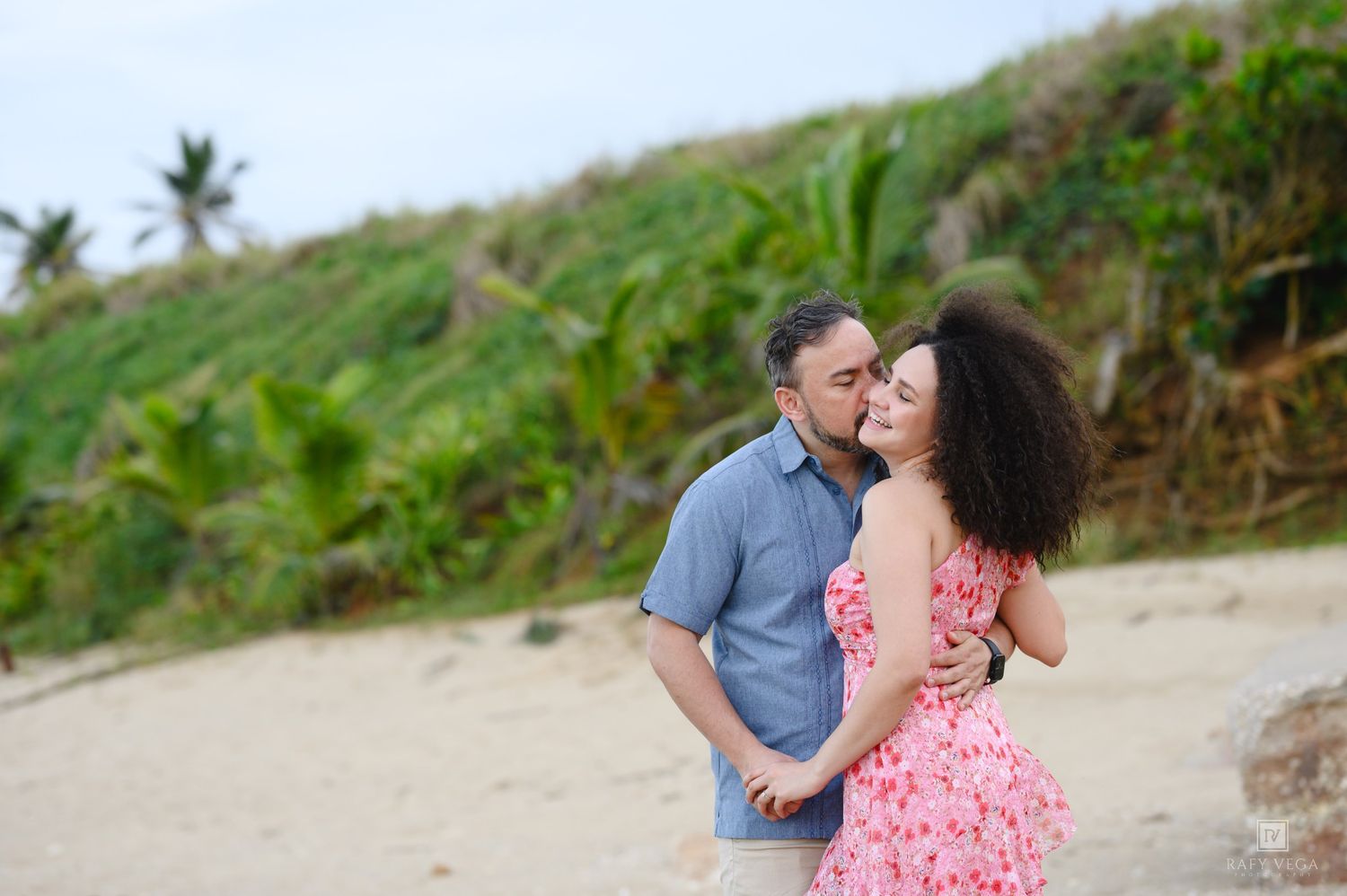 Couple sharing a romantic kiss on a sandy tropical beach surrounded by lush green palm trees.