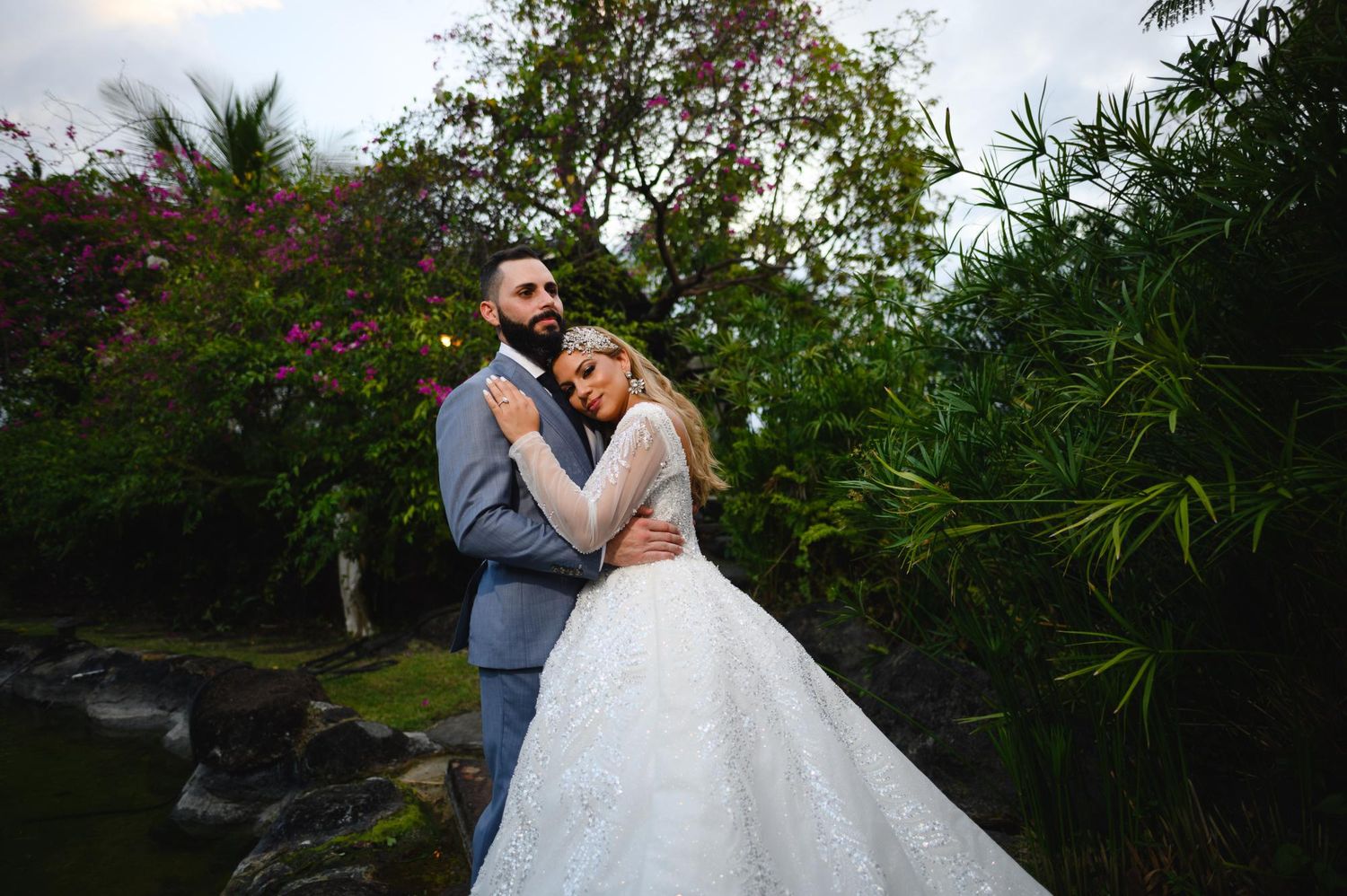 Bride and groom embrace in lush garden setting, she in white ball gown, he in gray suit, surrounded by tropical greenery.
