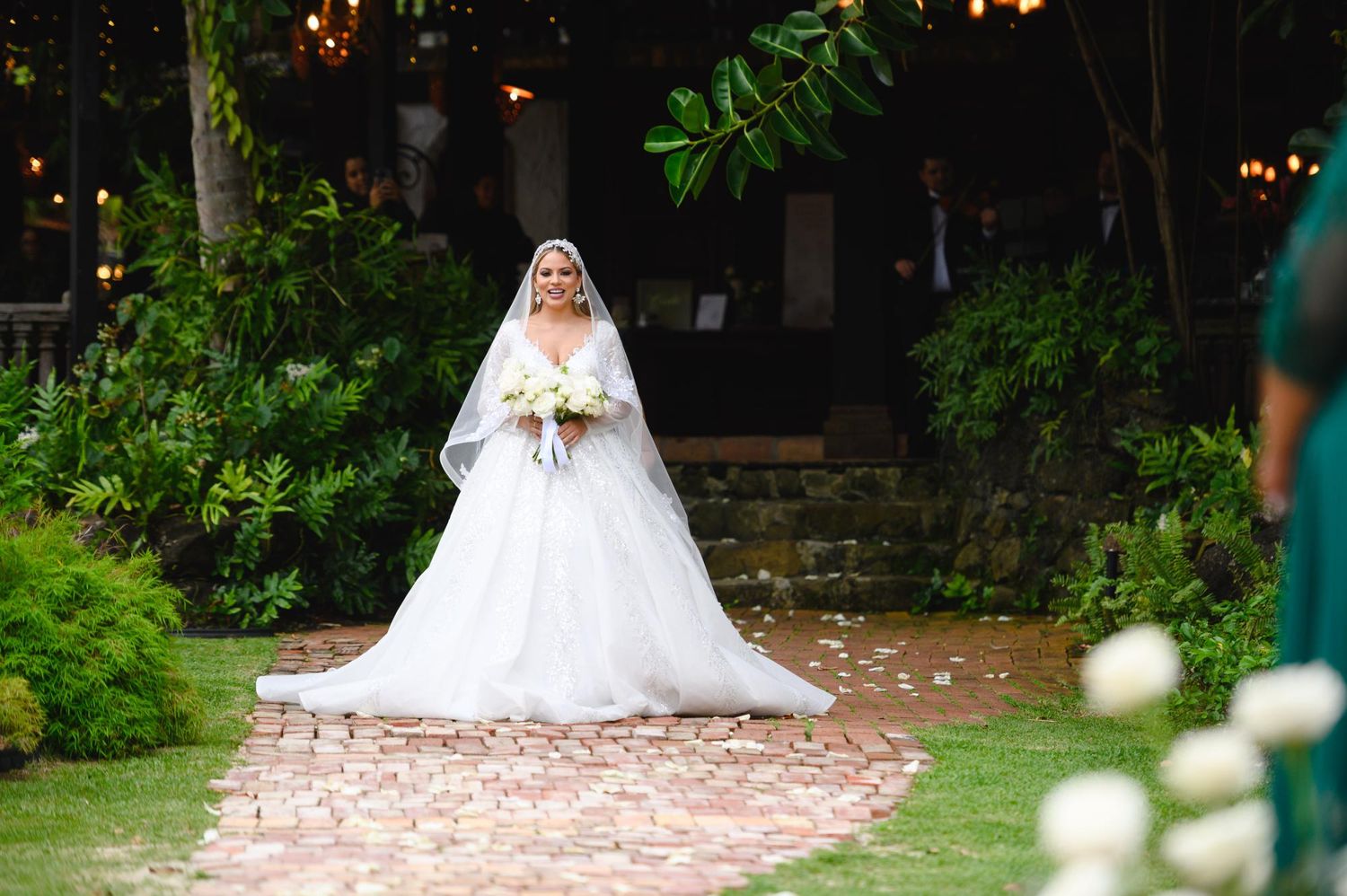 Bride in a white ball gown with veil walks down an outdoor garden aisle holding a bouquet at a wedding ceremony.