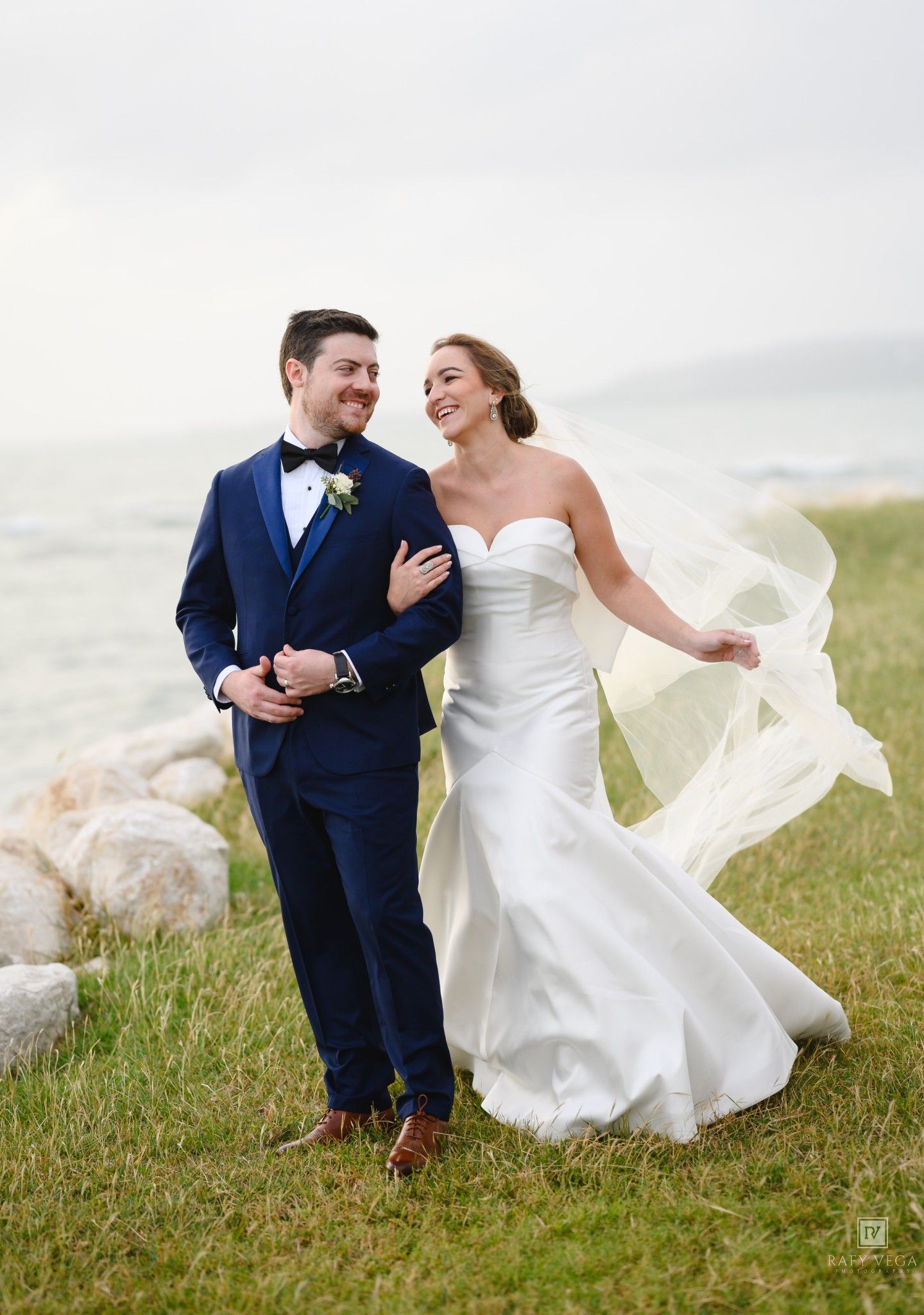 Bride and groom walking by the waterfront, her veil flowing in the wind, he wears a navy suit.