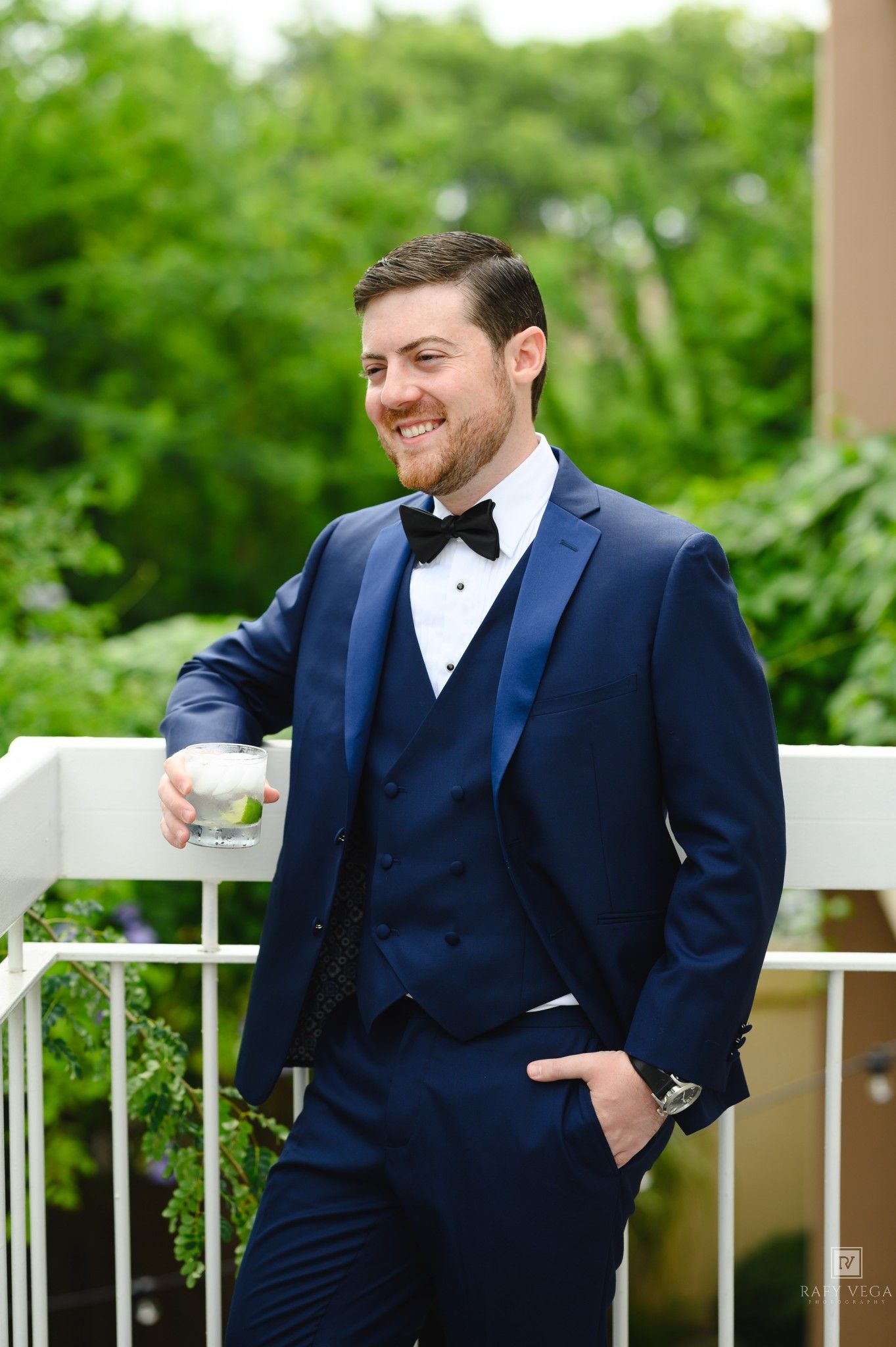 Smiling groom in navy blue three-piece tuxedo with black bow tie holding a drink on an outdoor balcony.