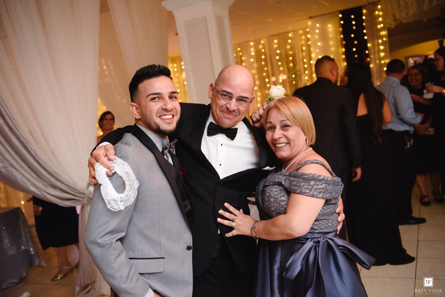 Three people smiling and posing together at a formal event, with fairy lights and elegant draping in the background.