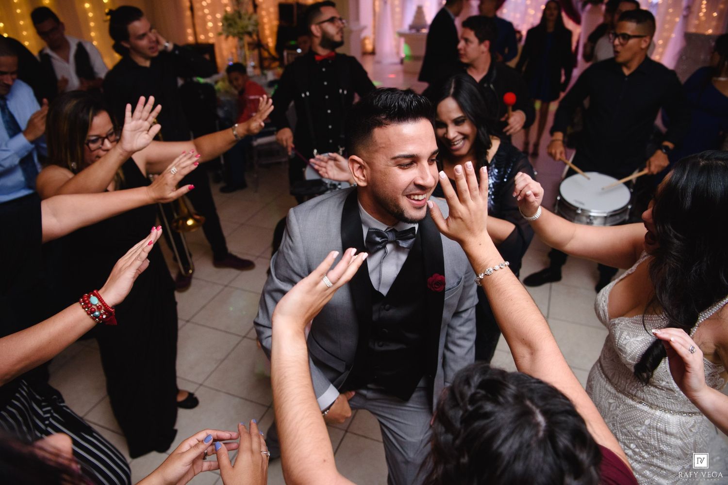 Groom in gray suit celebrated by guests on lively wedding reception dance floor.