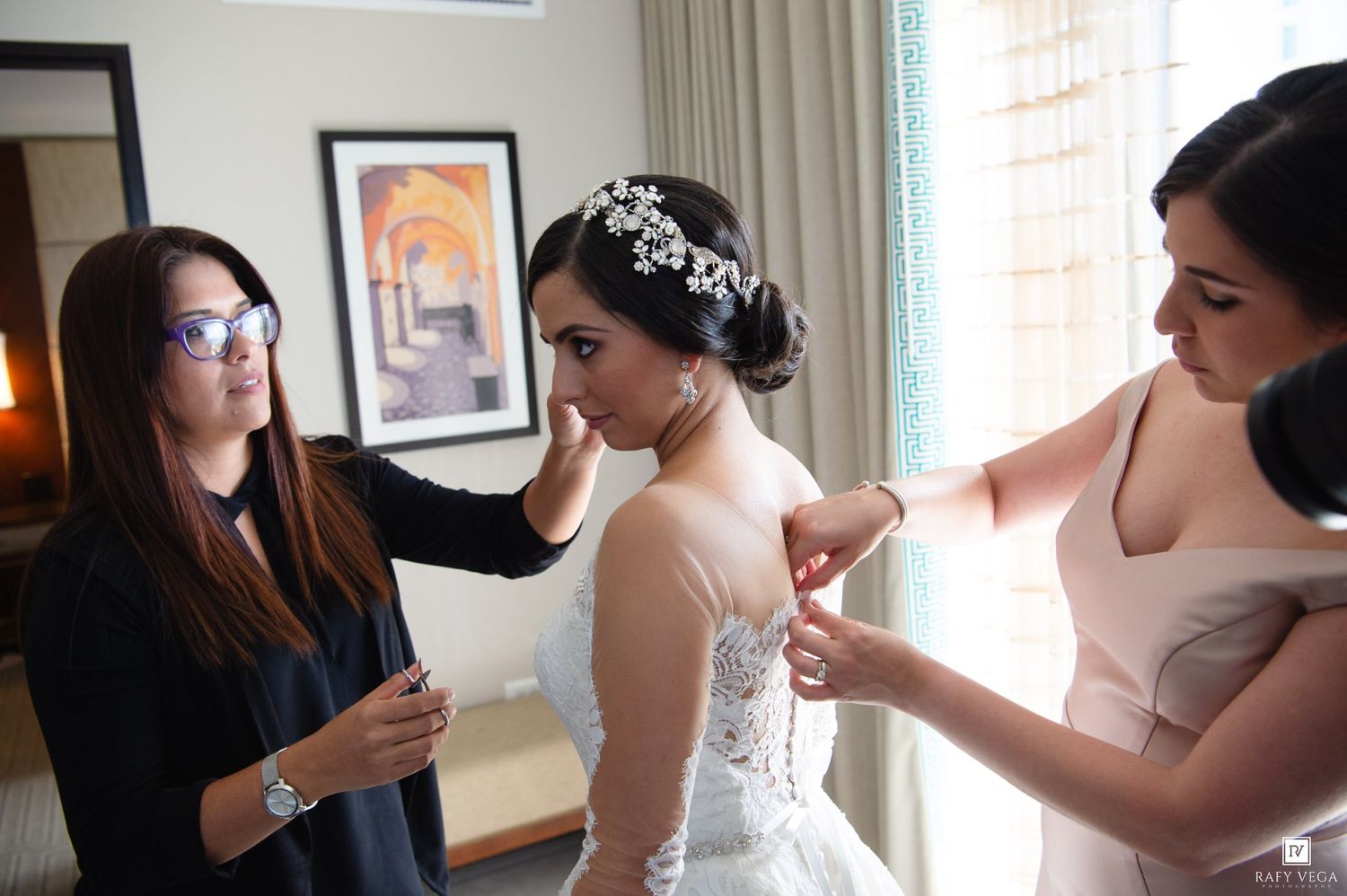 Bride in white lace wedding gown being helped by two women adjusting her dress and veil in a hotel room.