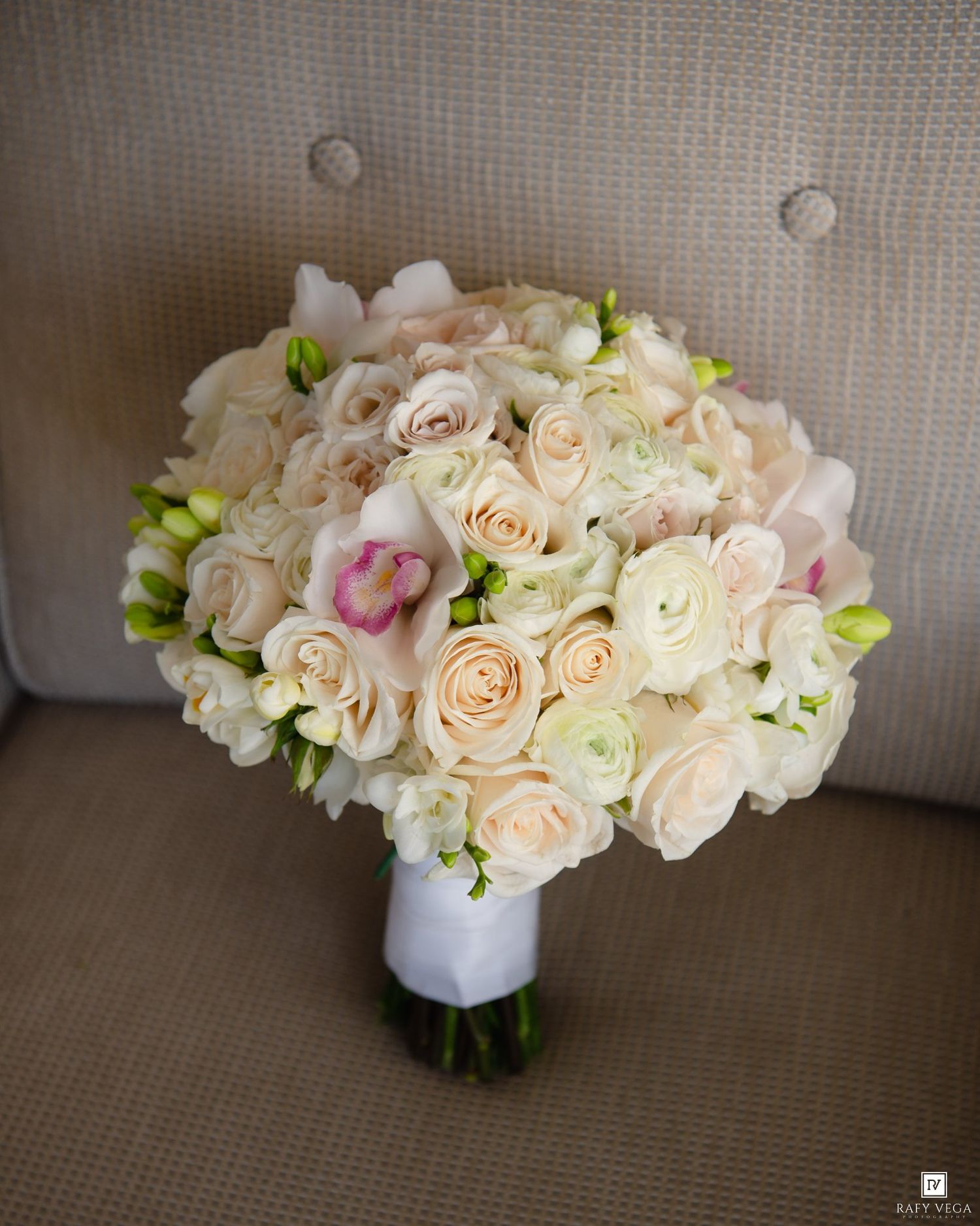 Elegant bridal bouquet of cream roses, white ranunculus, and pink orchids wrapped in white ribbon.
