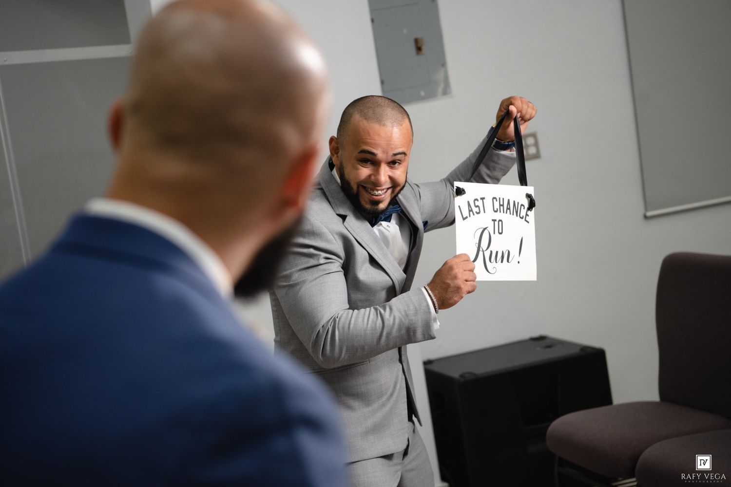 Groom in gray suit holds 'Last Chance to Run' sign while laughing with groomsman before wedding ceremony.