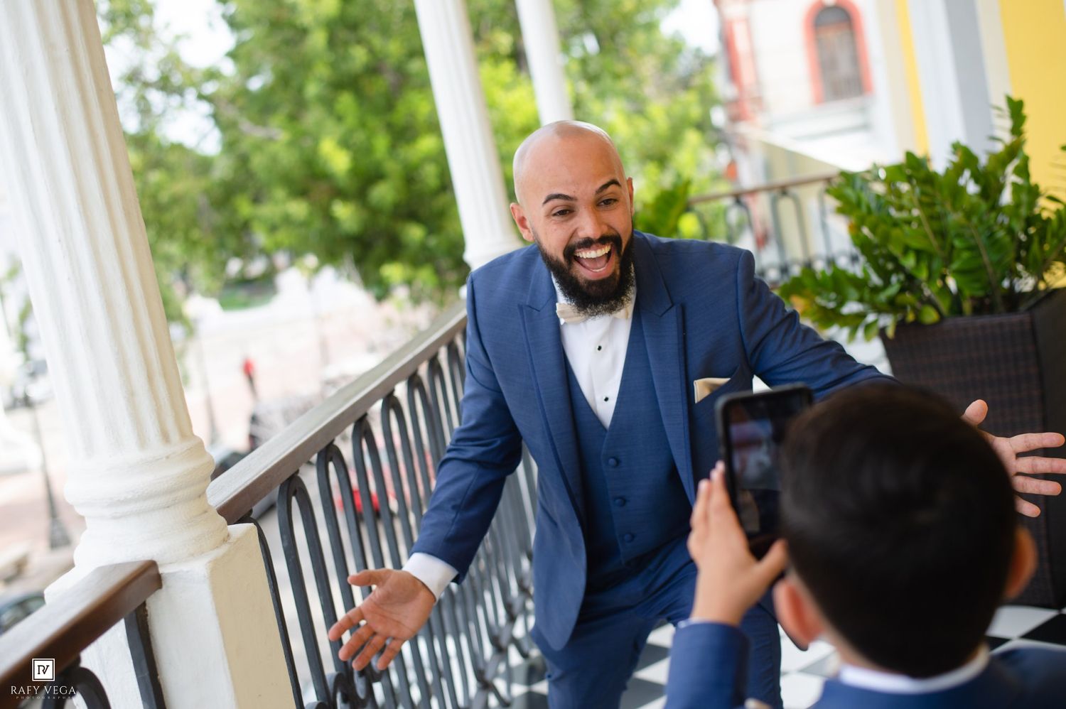 Man in blue suit smiling and posing for a photo on a balcony, being photographed by another person.