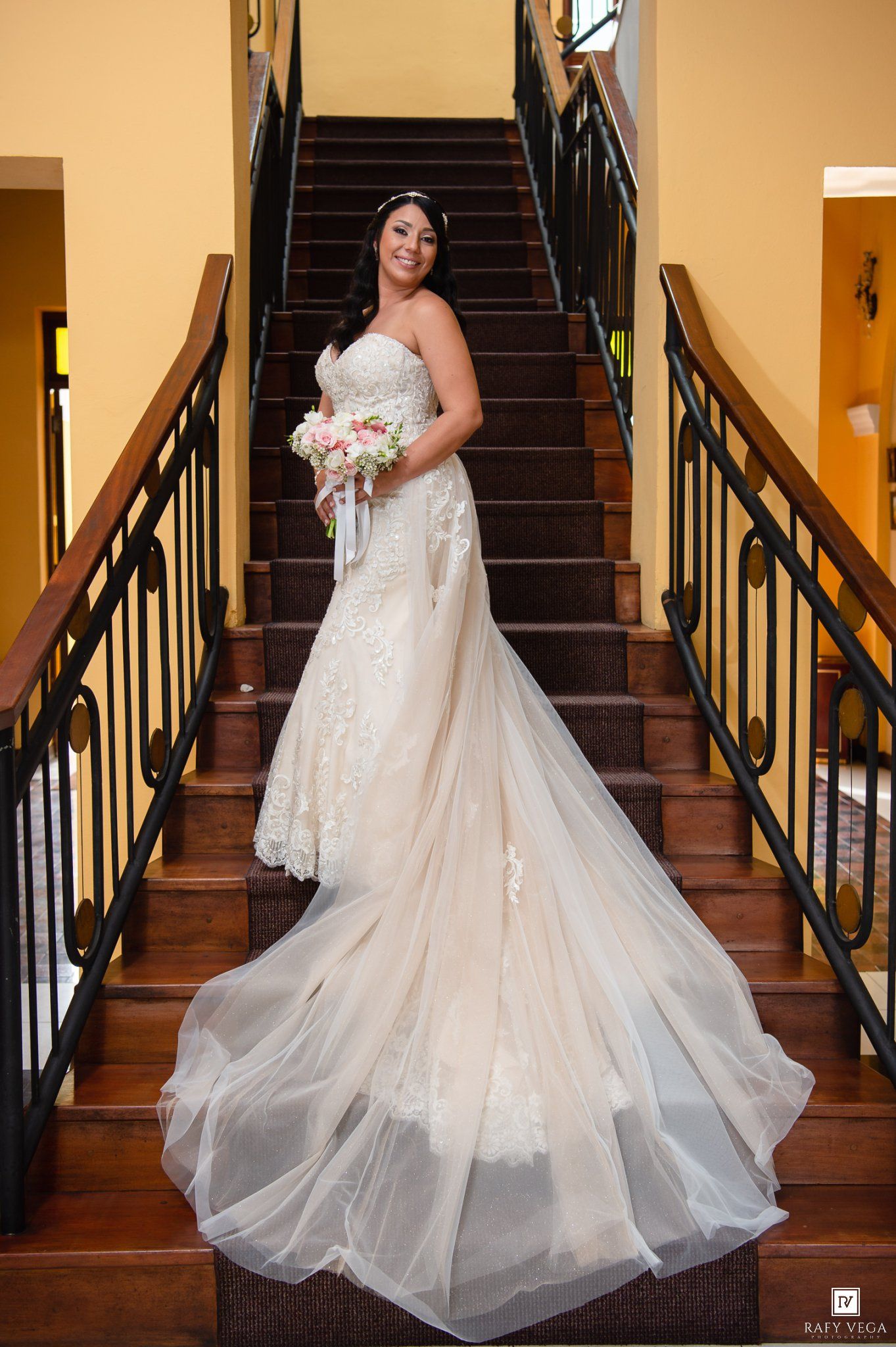 Bride in elegant lace wedding gown with long train holds bouquet on wooden staircase with black iron railing.