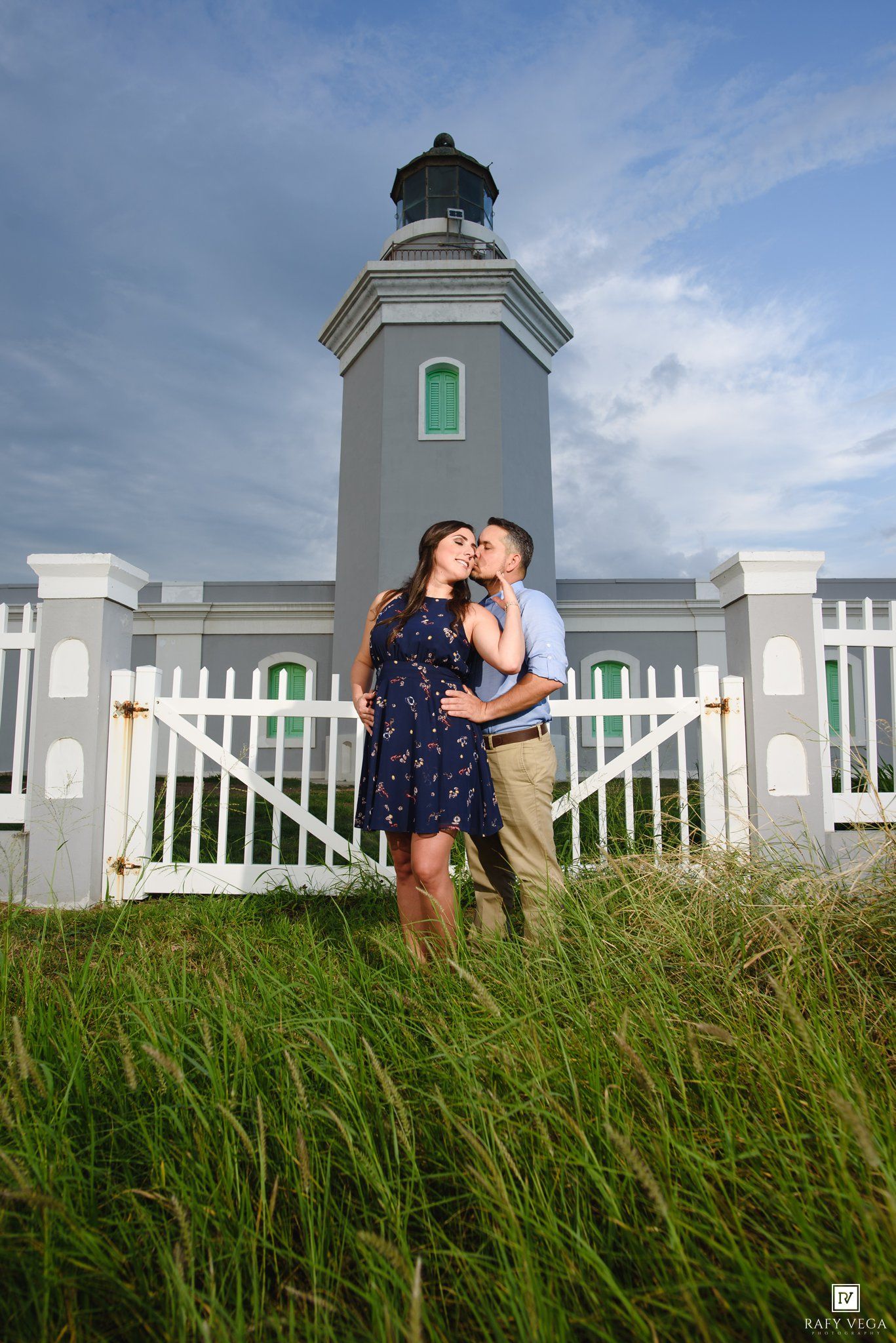 Couple sharing a romantic kiss in front of a gray lighthouse with white fence gate surrounded by lush green grass.