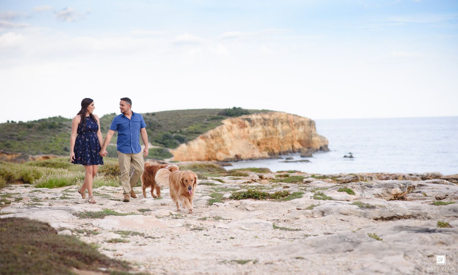 Couple walking golden retriever dog on rocky coastal cliffs with ocean and golden rock formations in background.
