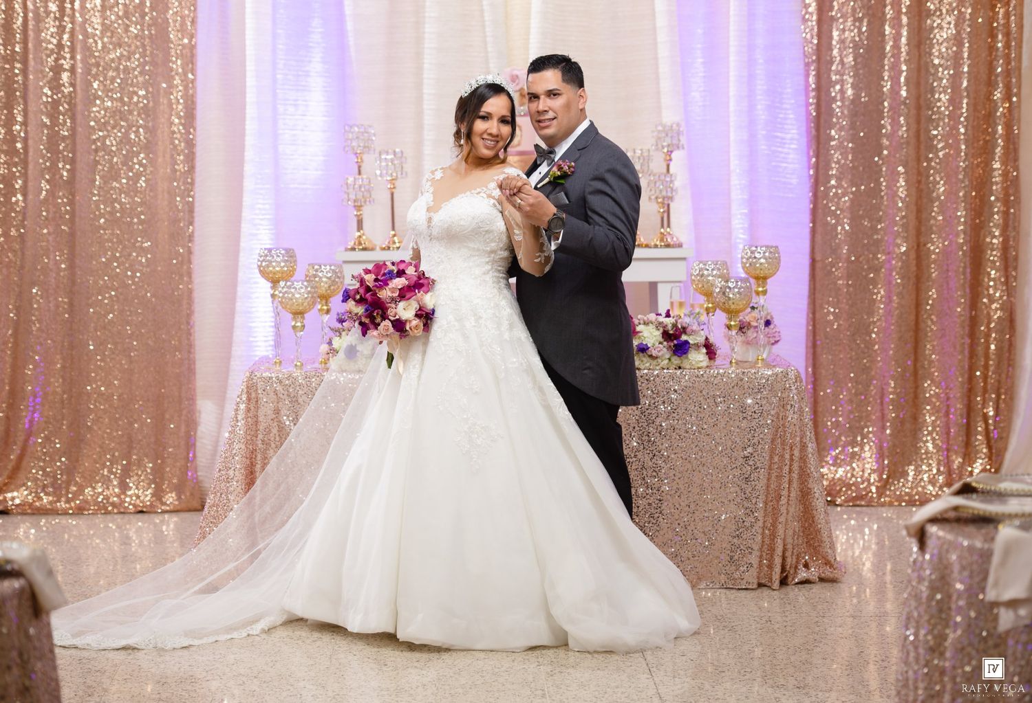 Bride and groom pose elegantly at their wedding reception with glittering rose gold sequin backdrop and floral arrangements.