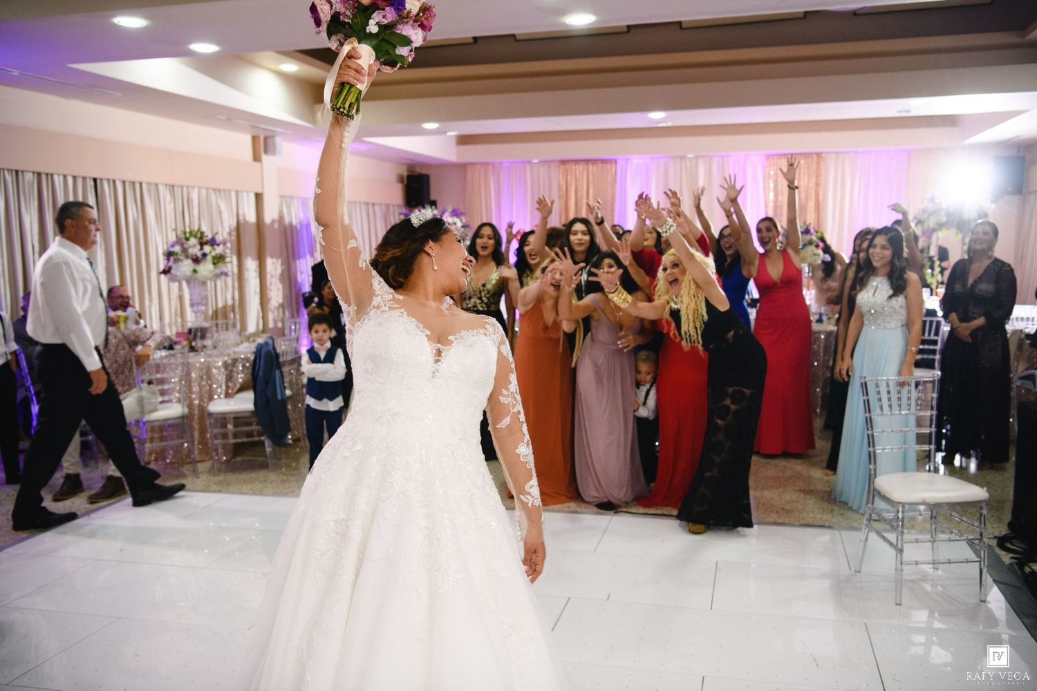 Bride in white lace gown tosses bouquet to excited guests on dance floor at elegant wedding reception.