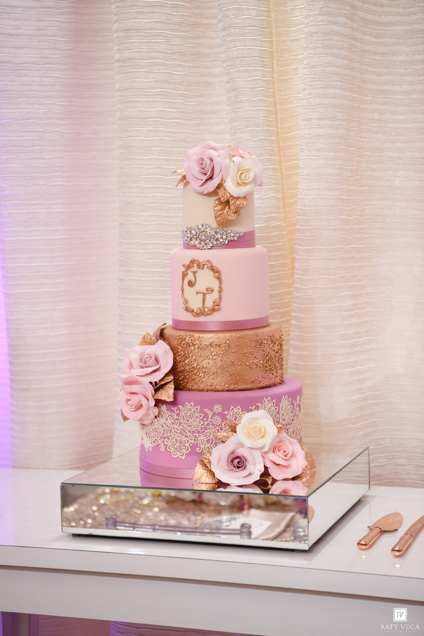 Elegant four-tier pink and gold wedding cake with sugar roses, lace details, and monogram on a mirrored base.