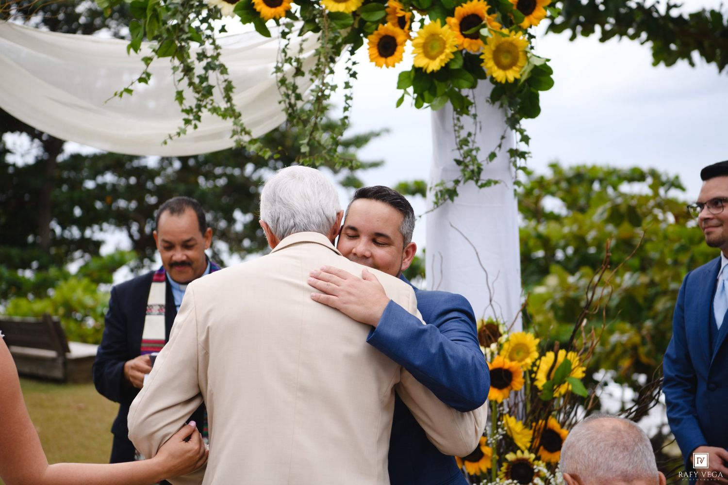 Groom in blue suit hugging guest at outdoor wedding ceremony decorated with sunflowers.