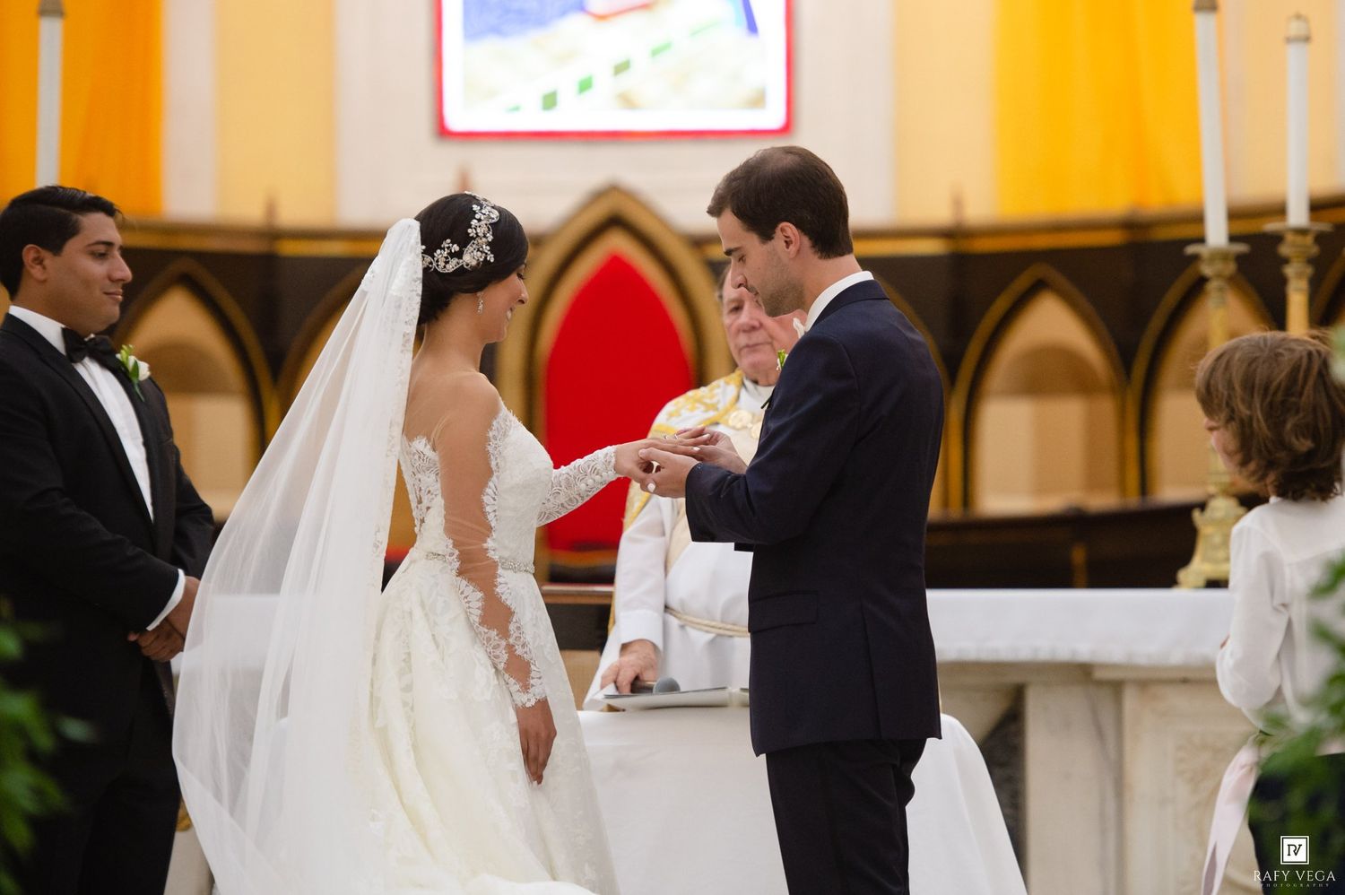 Groom places wedding ring on brides hand during romantic church ceremony with priest officiating at the altar.
