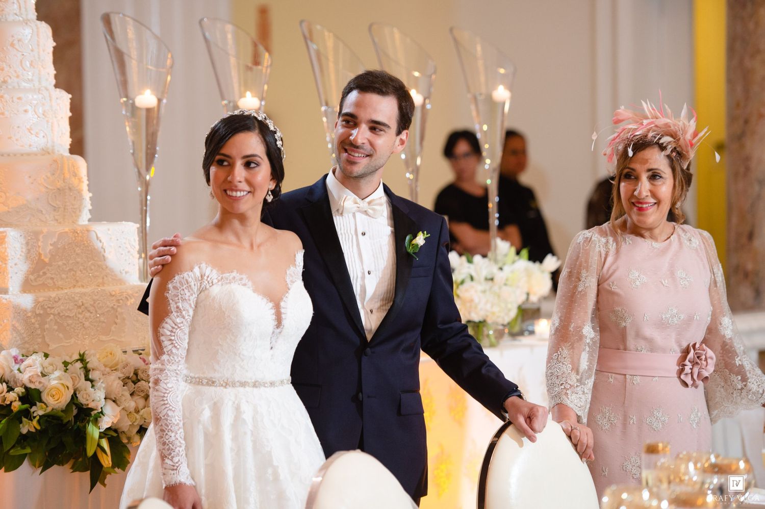Bride and groom smile with sparklers beside a tiered wedding cake at elegant reception.