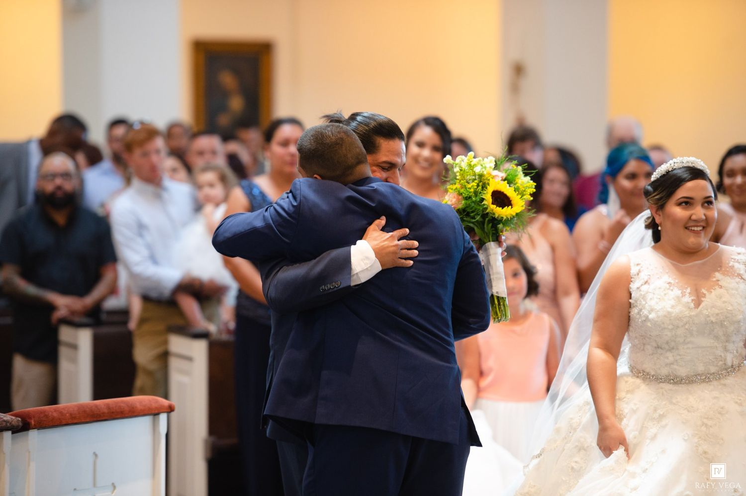 Groom embracing a guest at the altar during a church wedding ceremony, with the bride holding sunflowers nearby.