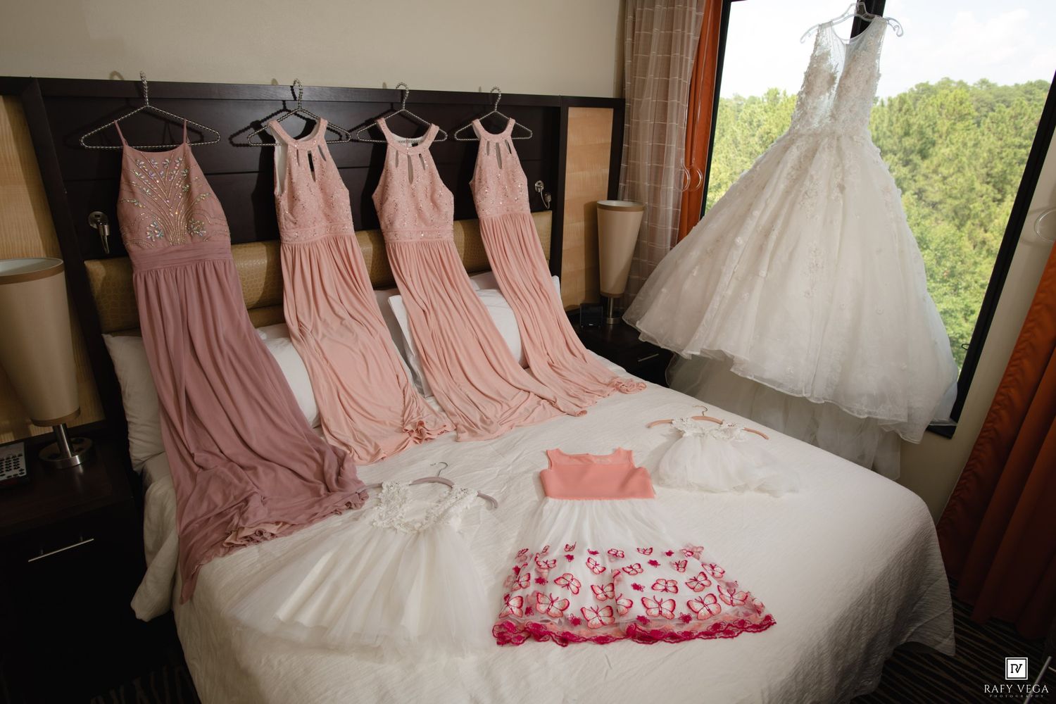 Pink bridesmaid dresses and white wedding gown hanging in hotel room with flower girl dress laid on bed.