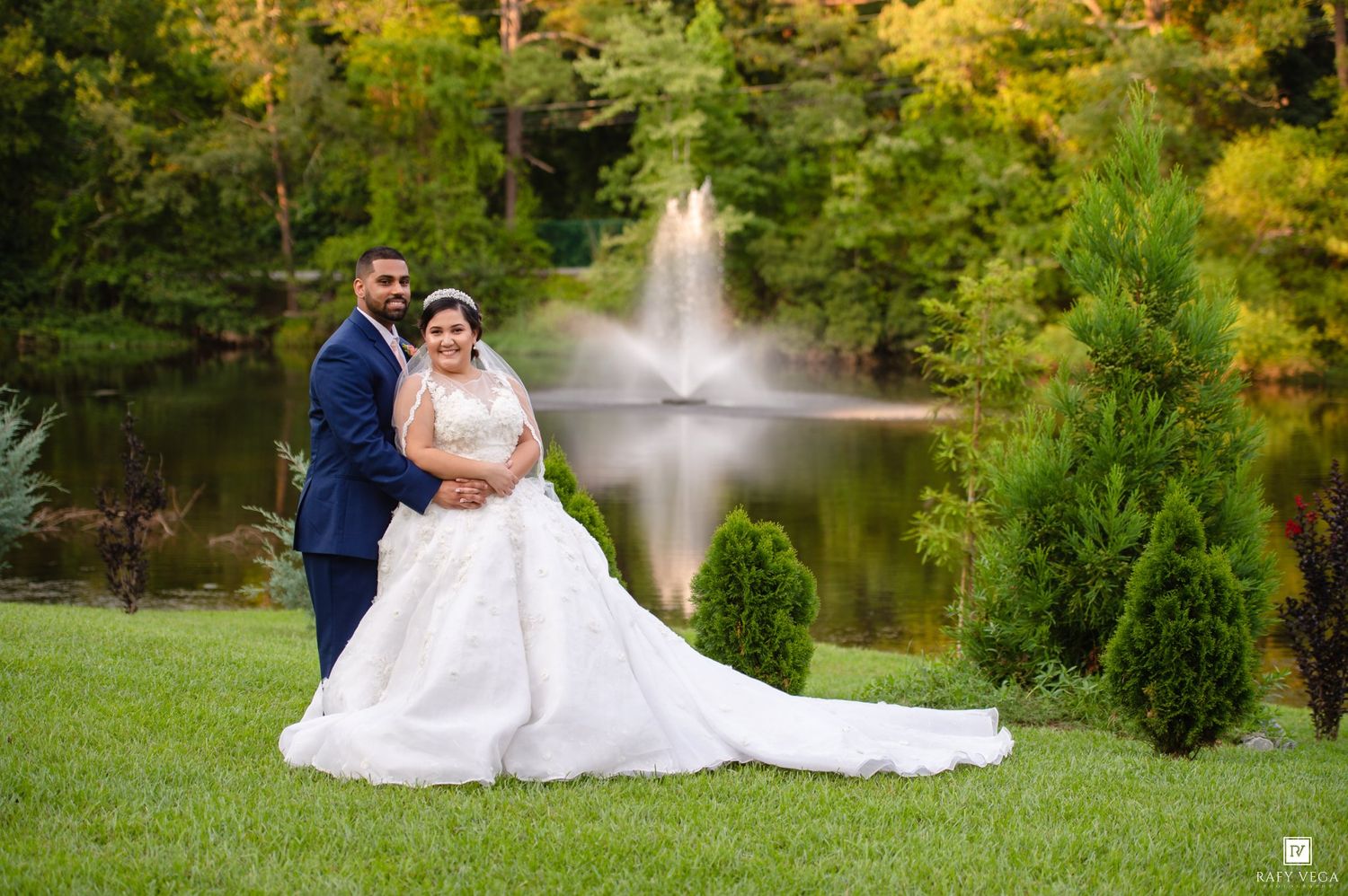 Bride and groom posing by a serene pond with a fountain and lush autumn trees in a romantic outdoor wedding setting.