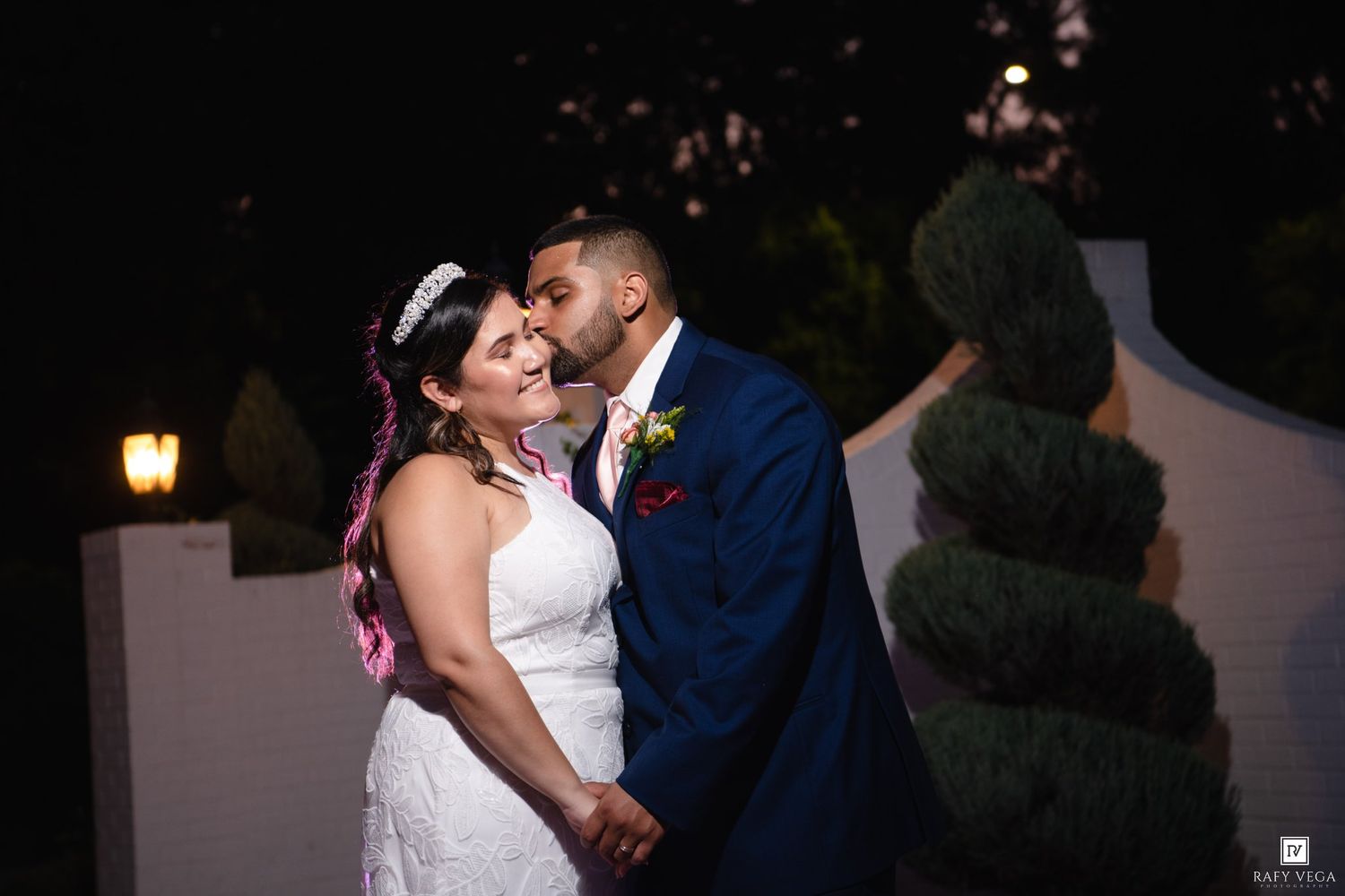 Bride and groom share a romantic kiss outdoors at night, with lush greenery and warm lights in the background.