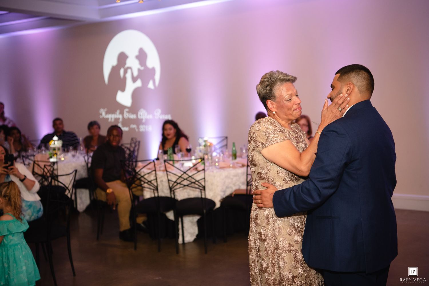 A groom shares an emotional mother-son dance at a wedding reception with soft purple lighting and guests seated in background.