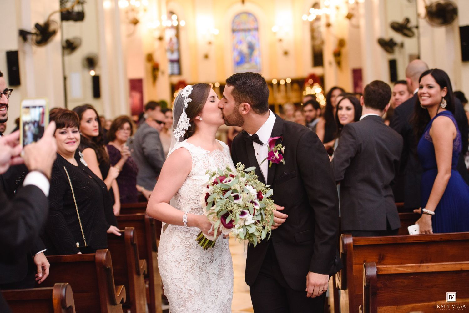 Newlywed couple sharing a first kiss at church altar, bride holding bouquet, guests celebrating in pews.