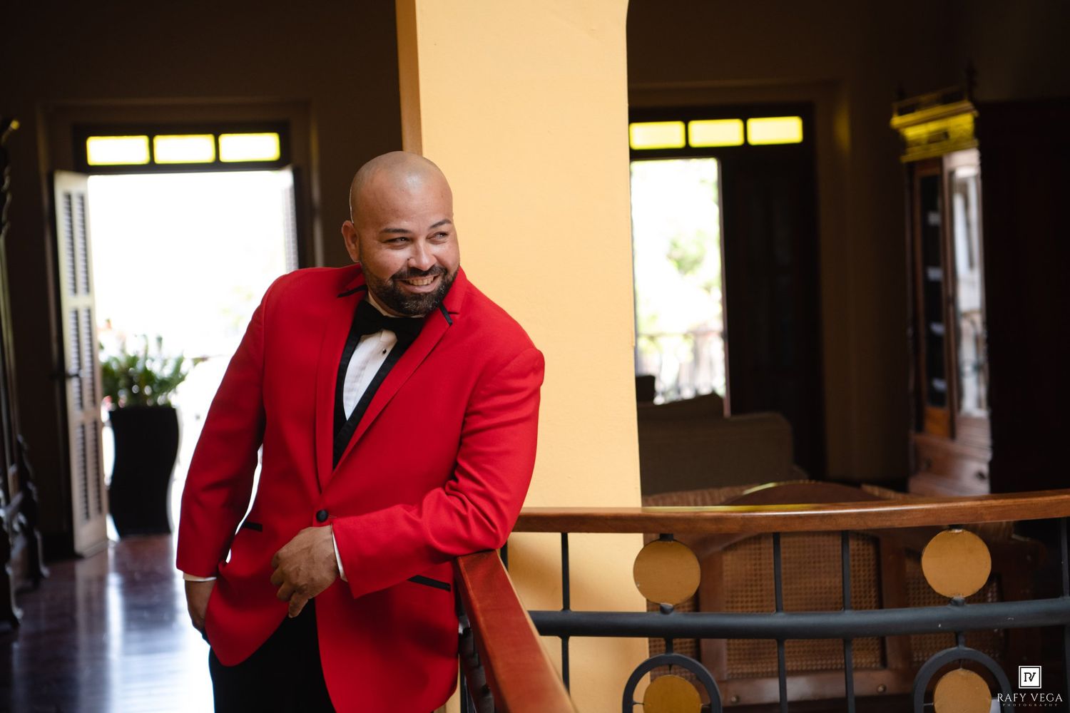 Man in red blazer and bow tie smiling while leaning on a railing in an elegant venue.