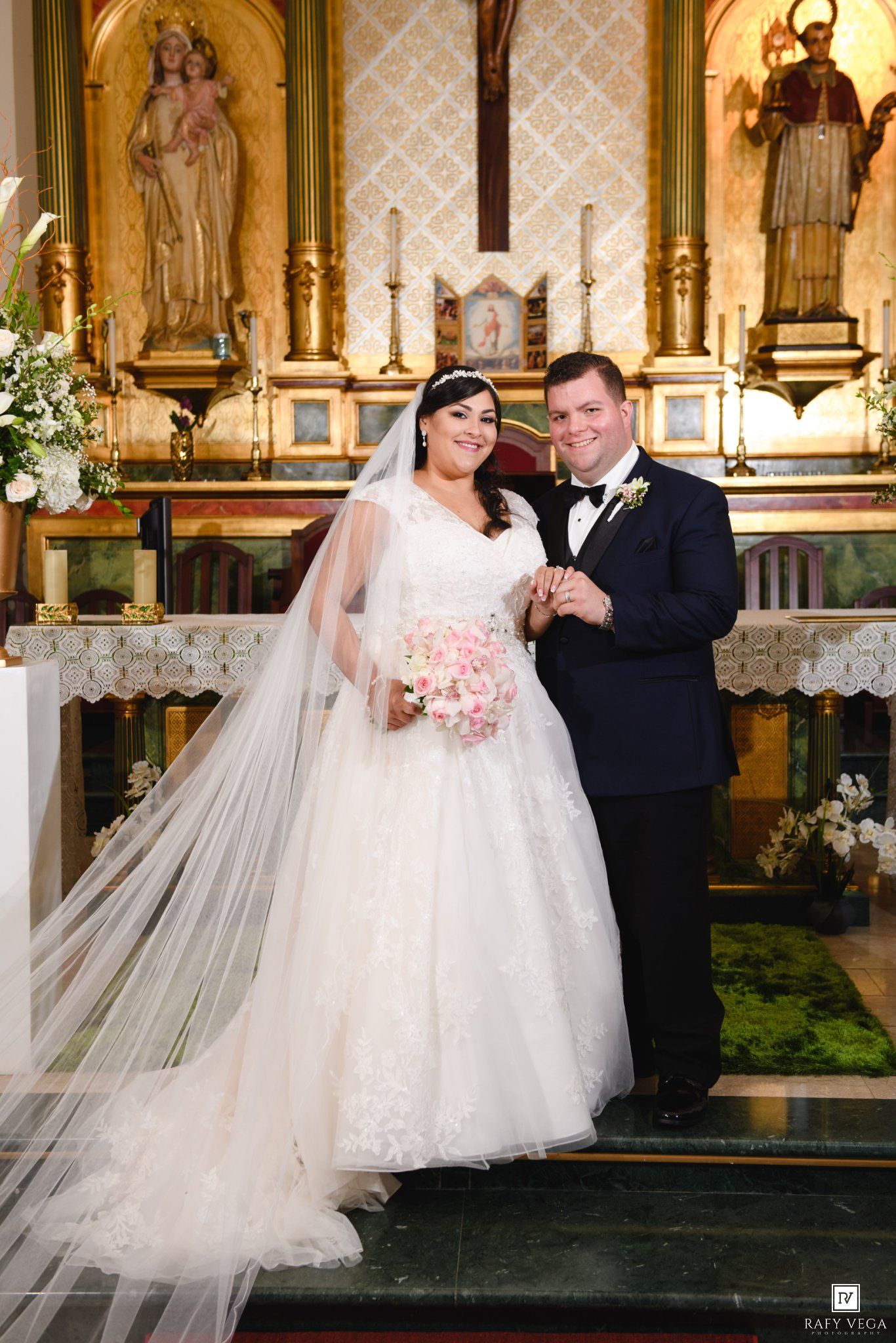 Bride and groom posing together at church altar, bride in white ballgown with veil holding pink bouquet.