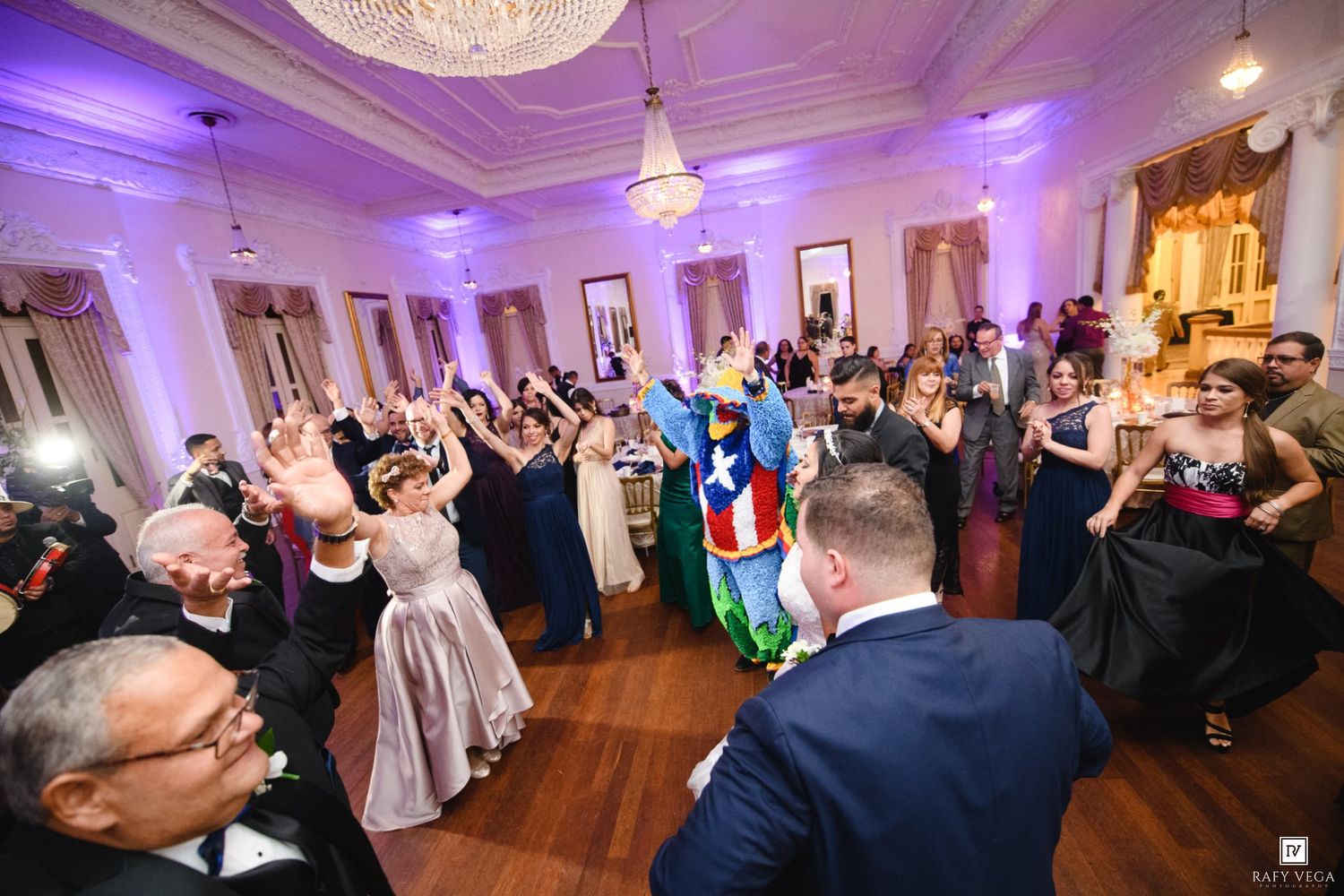 Guests dancing at an elegant wedding reception with purple lighting in a grand ballroom venue.