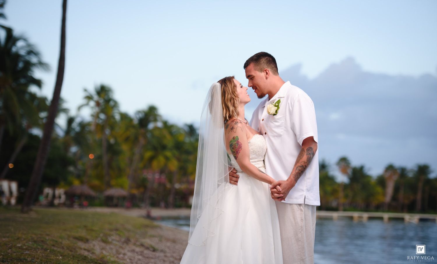 A bride and groom share a romantic moment on a tropical beach at sunset, surrounded by palm trees and calm blue water.