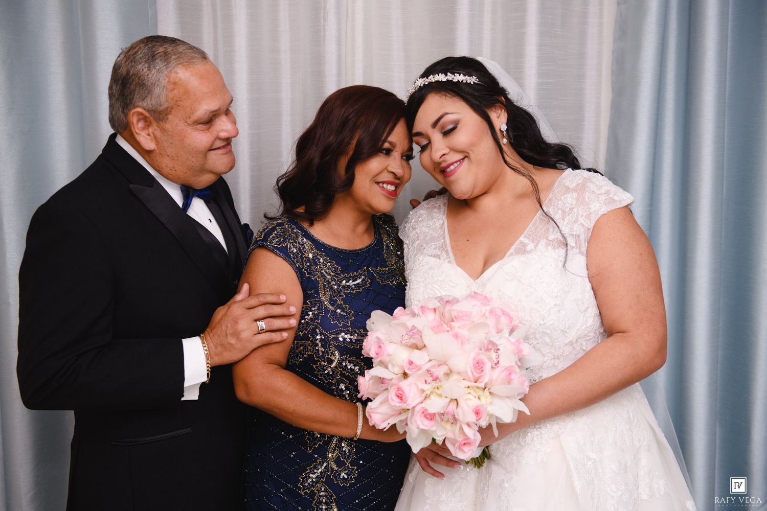 Bride in white lace gown holding pink bouquet poses with parents in formal attire at wedding celebration.