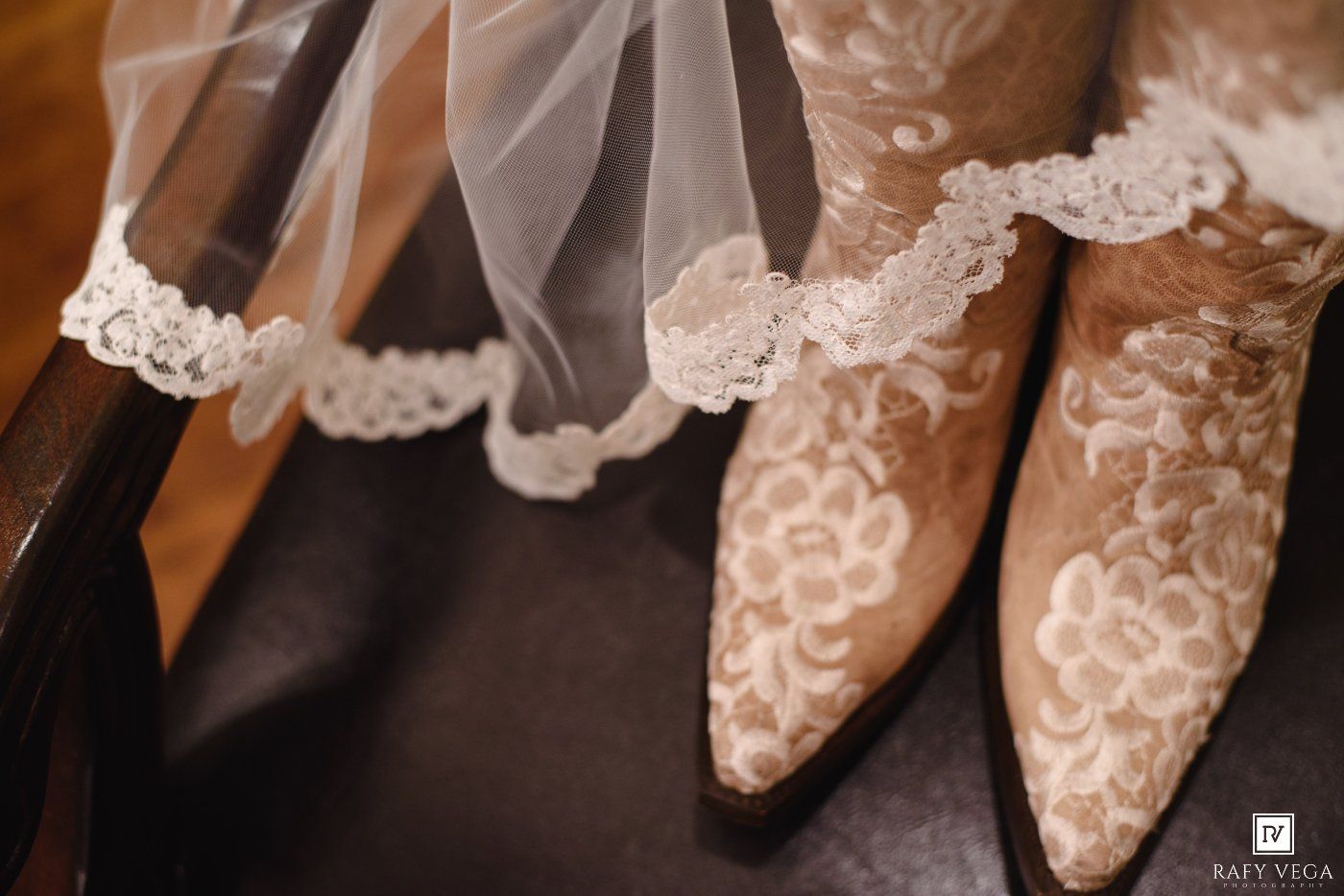 Elegant lace wedding veil draped over ornate floral embroidered bridal cowboy boots on a dark wooden chair.