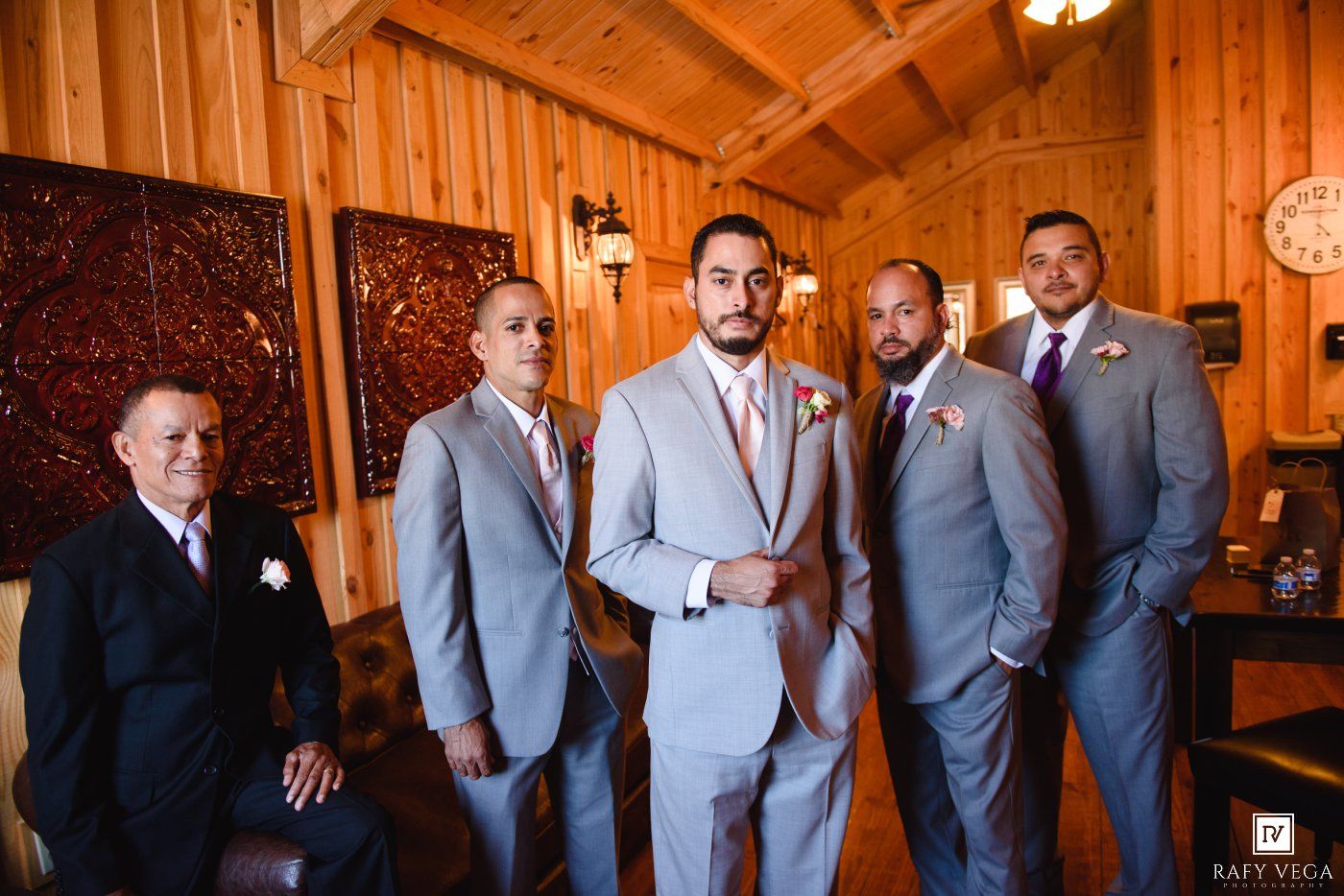 Groom and groomsmen in gray suits with pink ties pose in a rustic wooden venue before a wedding ceremony.