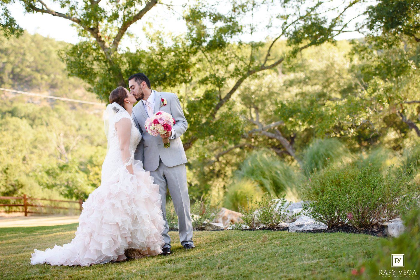 Bride and groom share a romantic kiss outdoors on green grass surrounded by lush trees at a beautiful garden wedding.