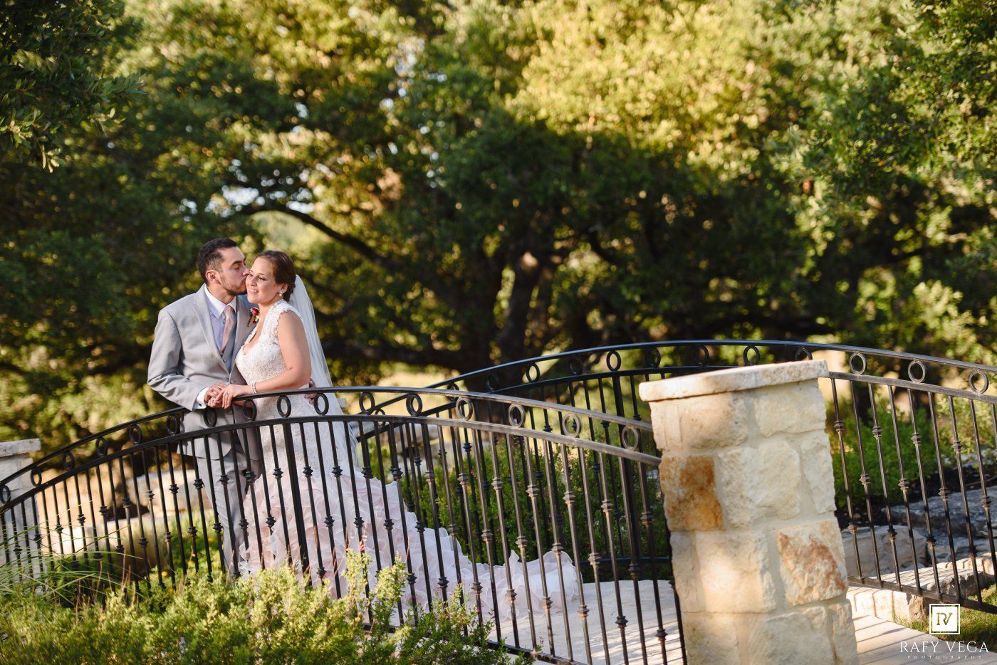 A couple shares a romantic moment on a stone bridge surrounded by lush green trees on their wedding day.