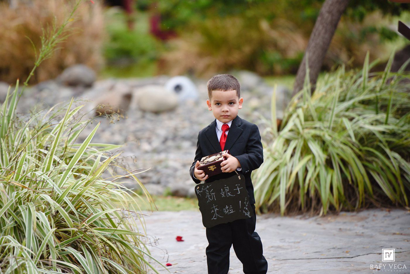 Young ring bearer in black suit and red tie holds decorative box at wedding.