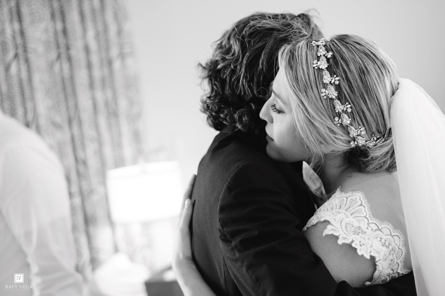 Bride in lace dress and floral headpiece shares tender embrace in black and white photo.