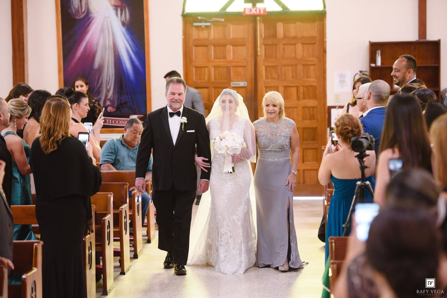 Bride walks down church aisle with groom and bridesmaid during wedding ceremony, guests seated in wooden pews.