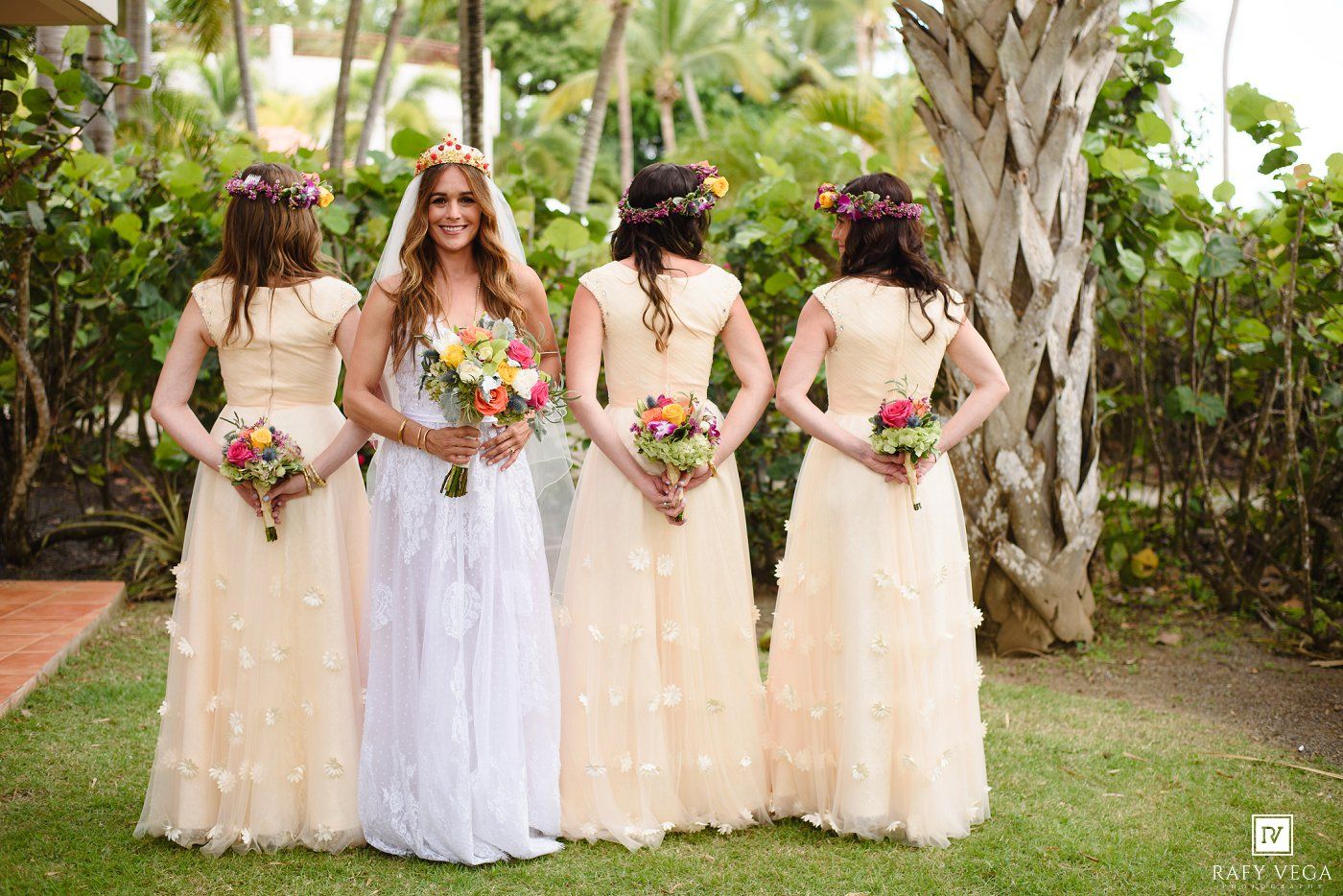 Bride and three bridesmaids in cream dresses with floral crowns holding colorful bouquets at an outdoor tropical wedding.