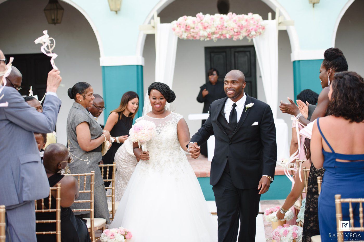 Bride and groom walk down the aisle at an elegant outdoor wedding ceremony with floral arch and guests cheering.