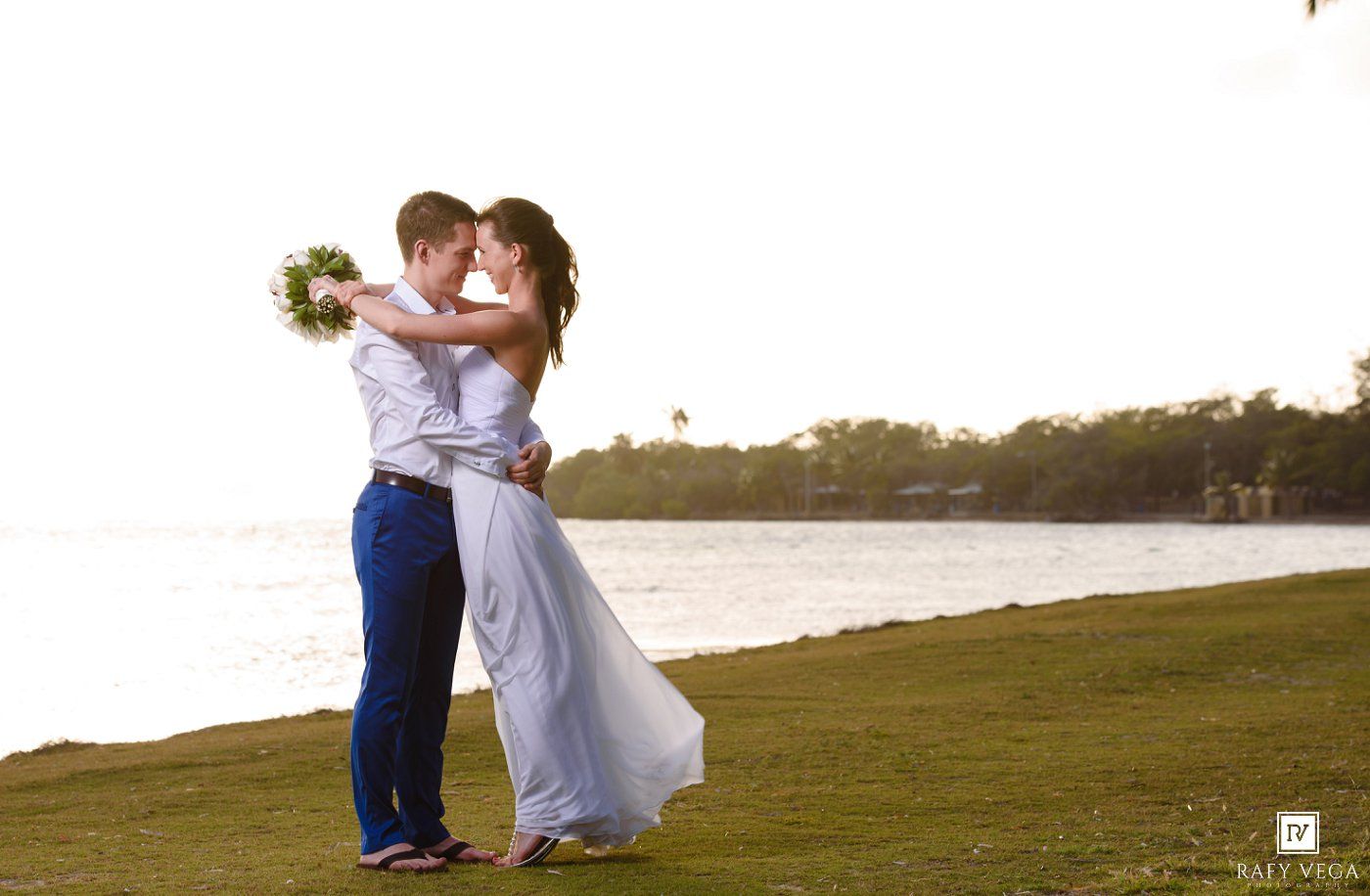 Couple sharing a romantic kiss in wedding attire on a lush green lawn near a serene waterfront at sunset.