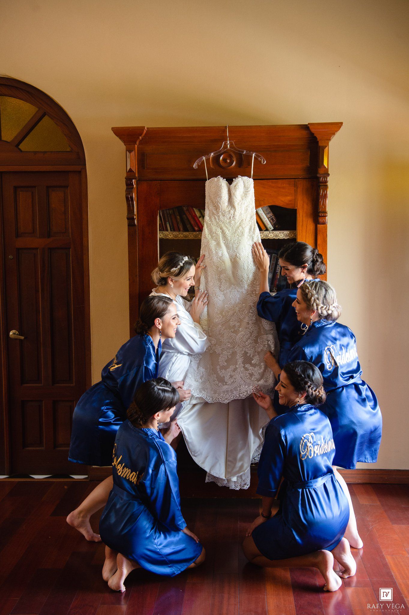 Bridesmaids in blue satin robes admire a lace wedding dress hanging on a wooden armoire before the ceremony.