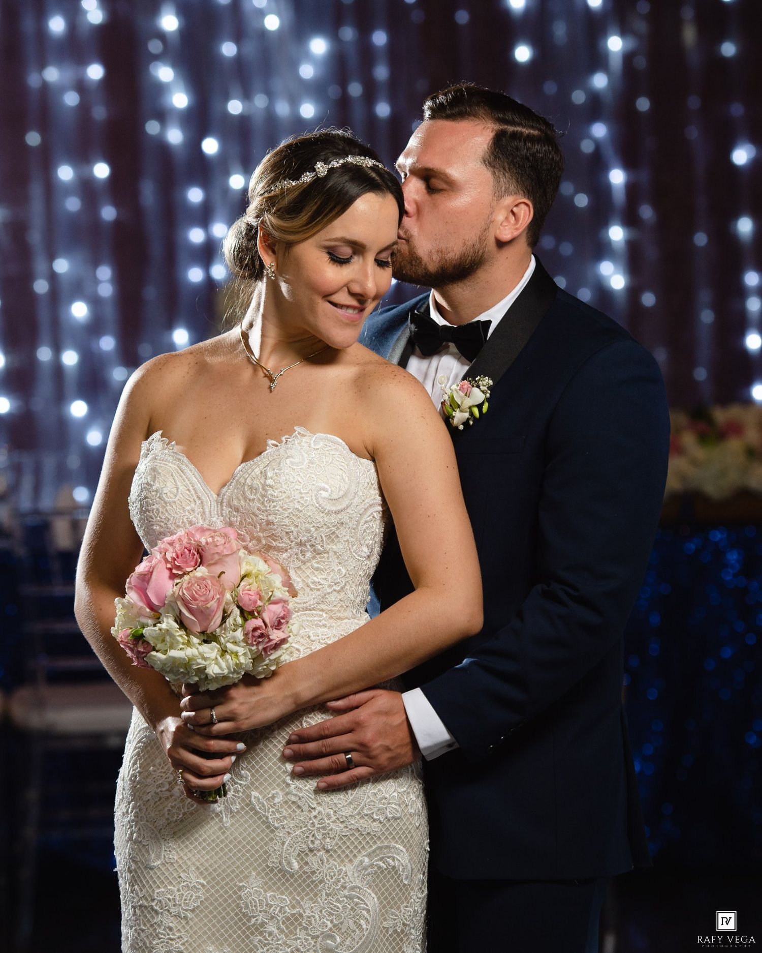 Groom kisses bride on cheek as she holds a pink rose bouquet before a sparkling fairy light curtain backdrop.
