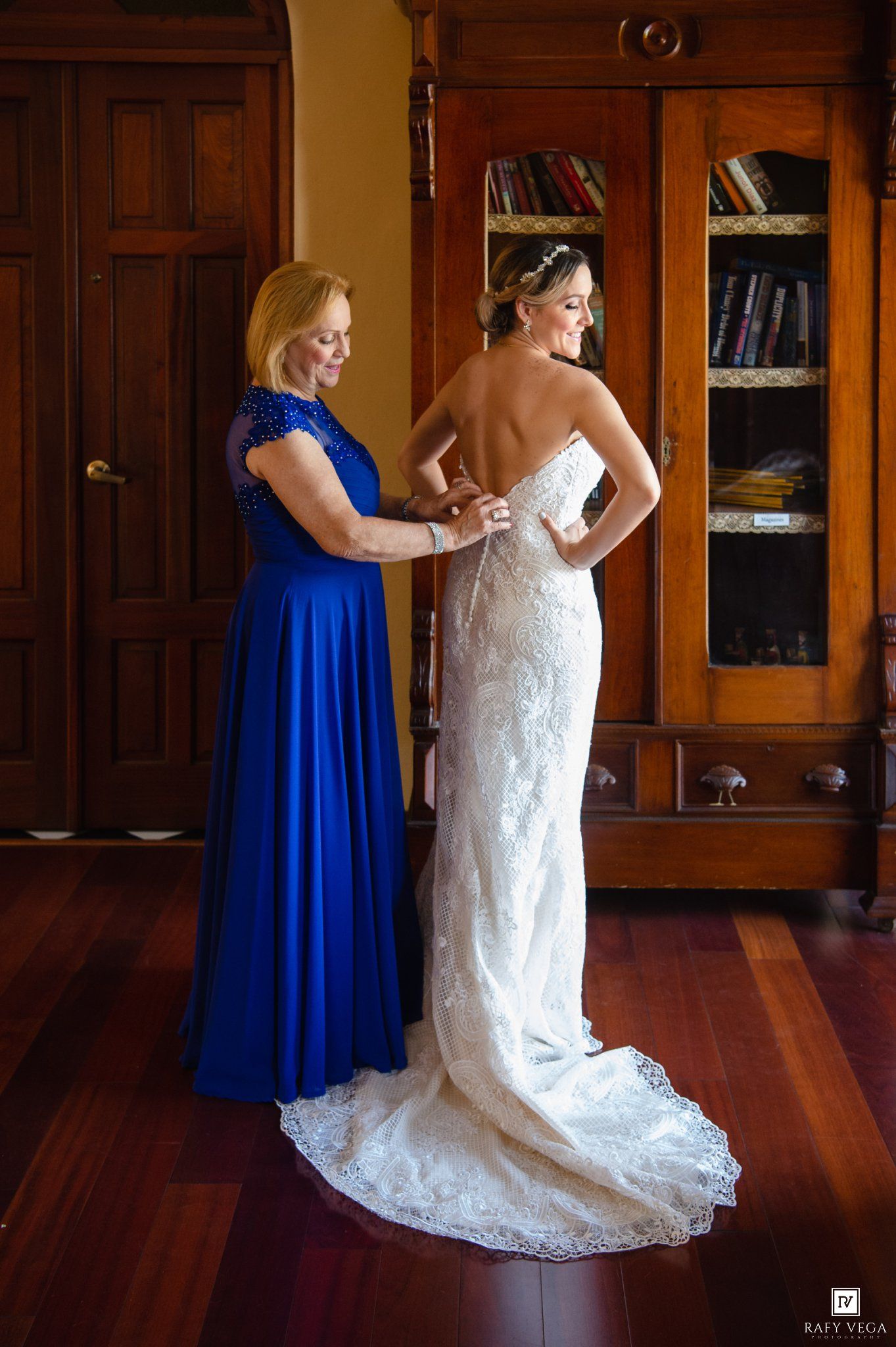 A bride in white lace gown being helped by a woman in royal blue dress in an elegant wood-paneled room.