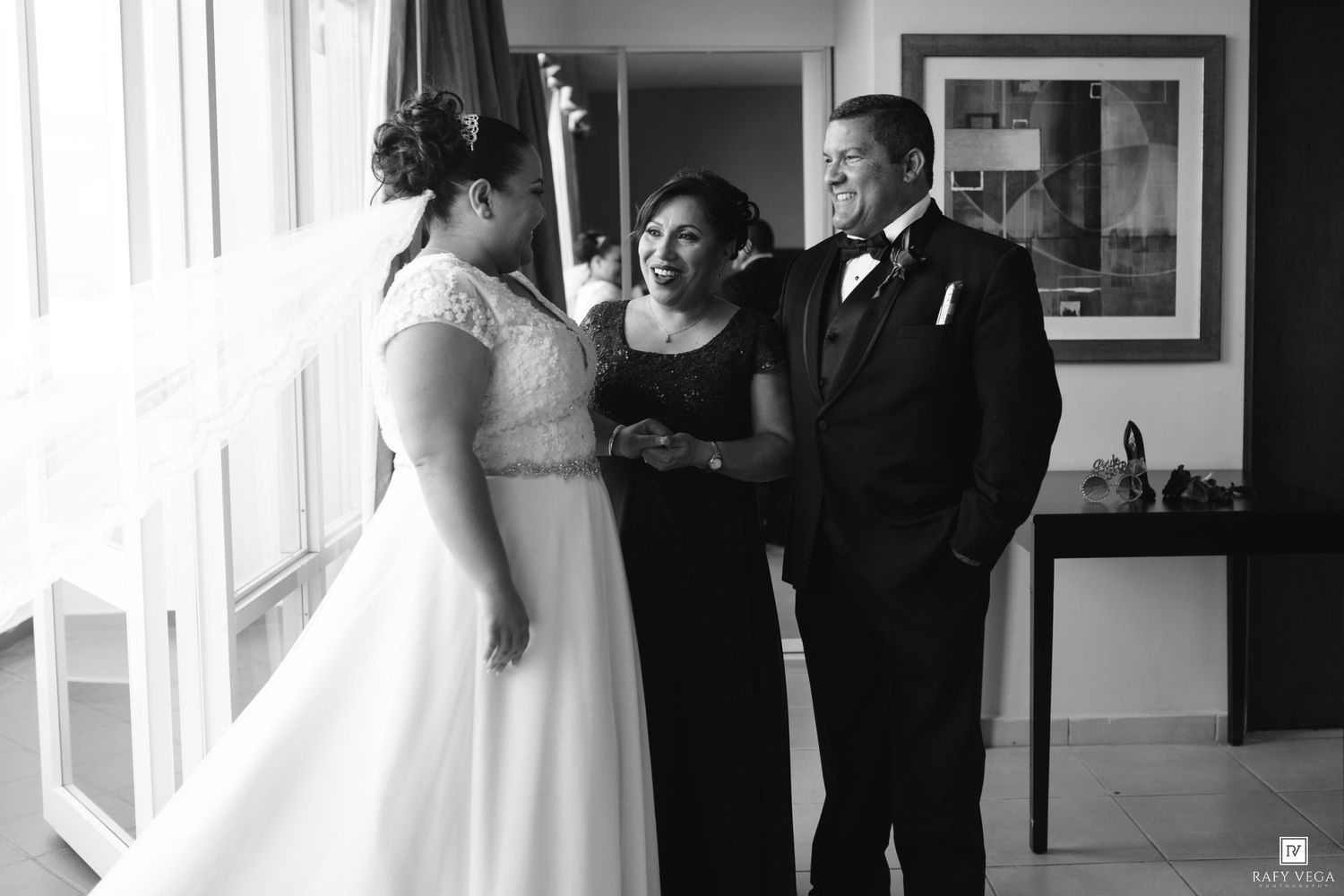 Black and white wedding photo of bride in white gown with two guests smiling near large windows.