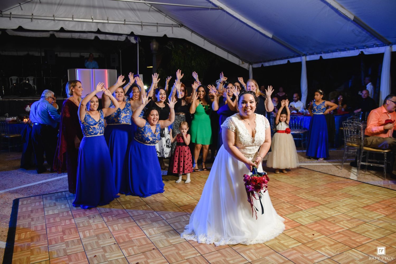 Bride tossing bouquet to excited guests in blue dresses at an outdoor wedding reception.