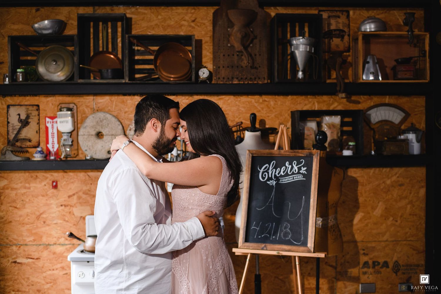 Couple sharing a romantic embrace in a rustic kitchen setting with a Cheers chalkboard sign.