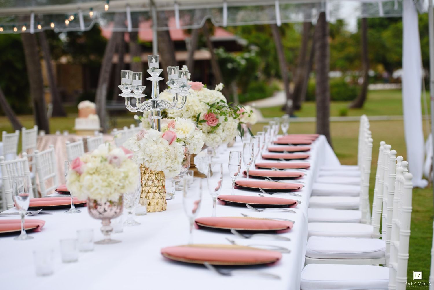 Elegant outdoor wedding reception table with white chairs, pink plates, floral centerpieces, and candelabras on a white tablecloth.