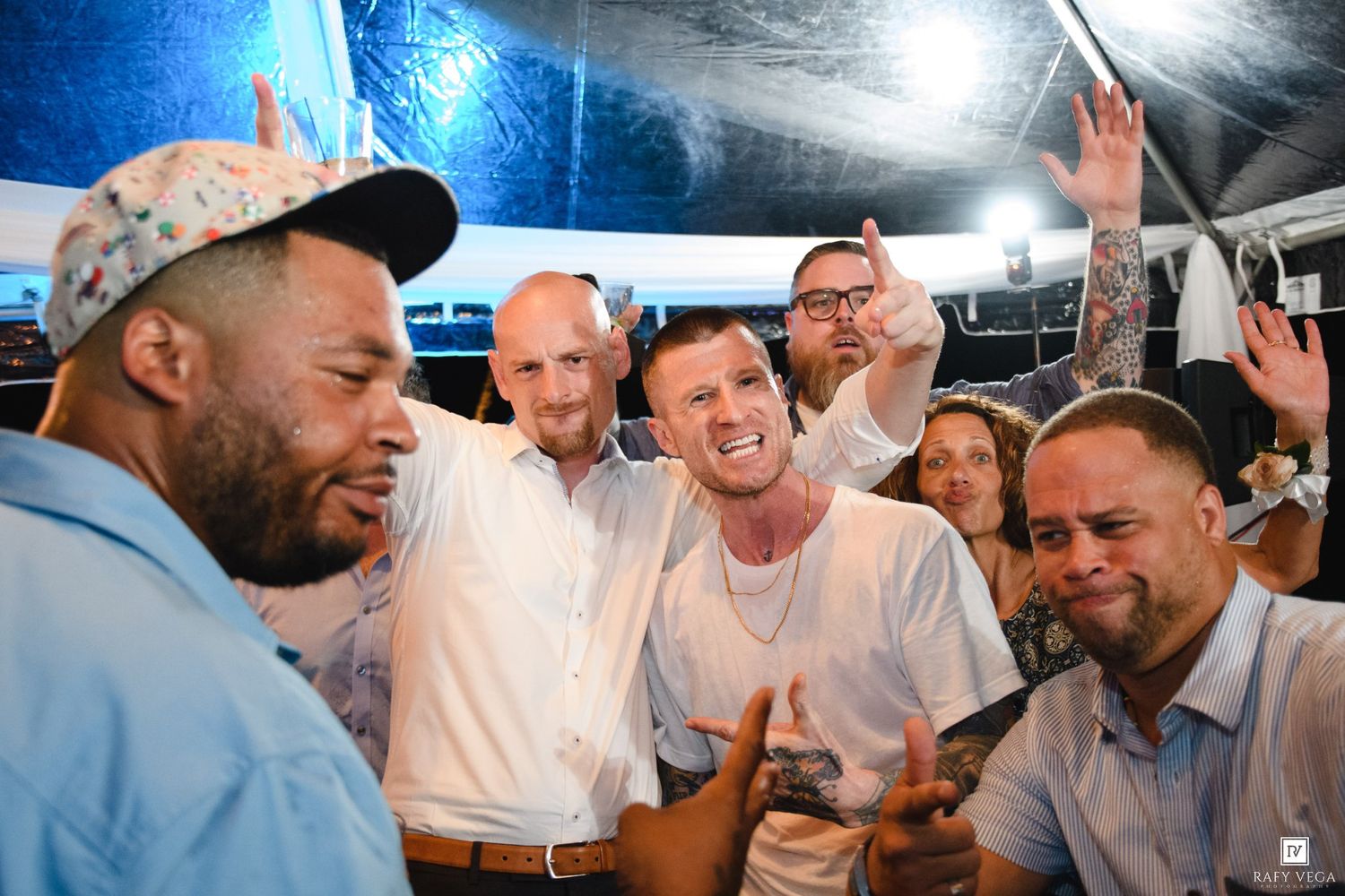Group of men celebrating at an outdoor party event under a tent, smiling and raising hands in excitement.